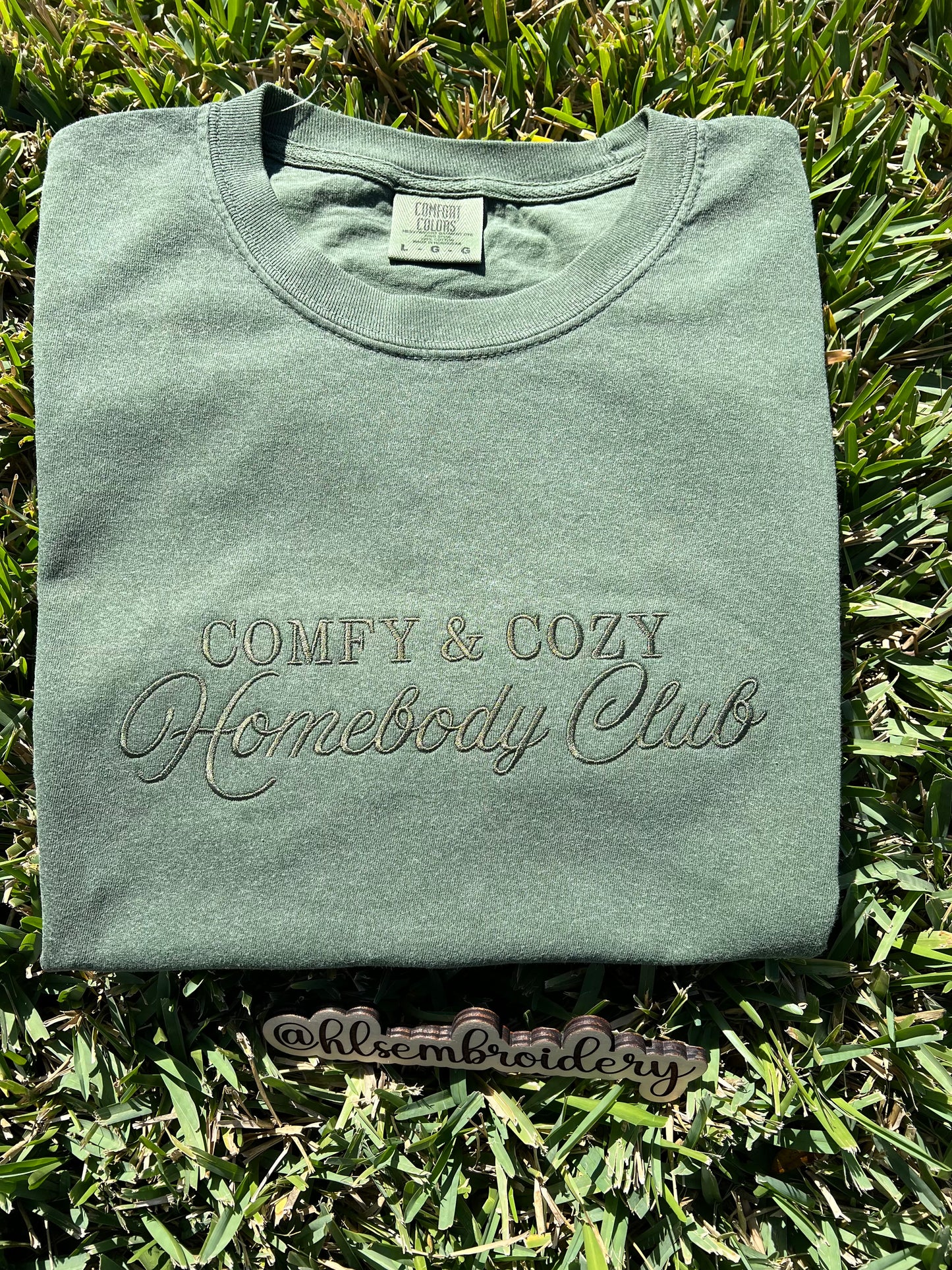 Folded green sweatshirt with 'Comfy & Cozy Homebody Club' text on grass