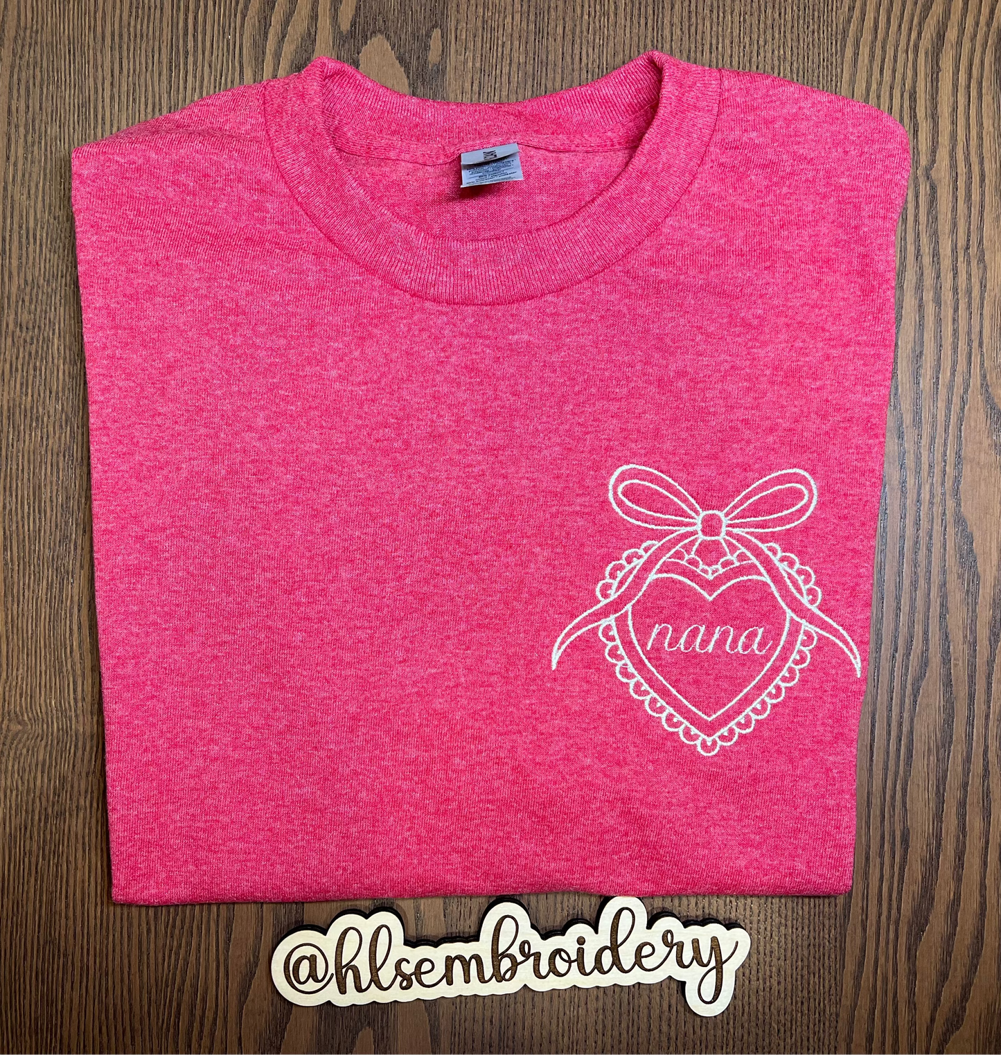 Folded pink t-shirt with white embroidery design on a wooden surface, featuring the brand 'hlsembroidery'.