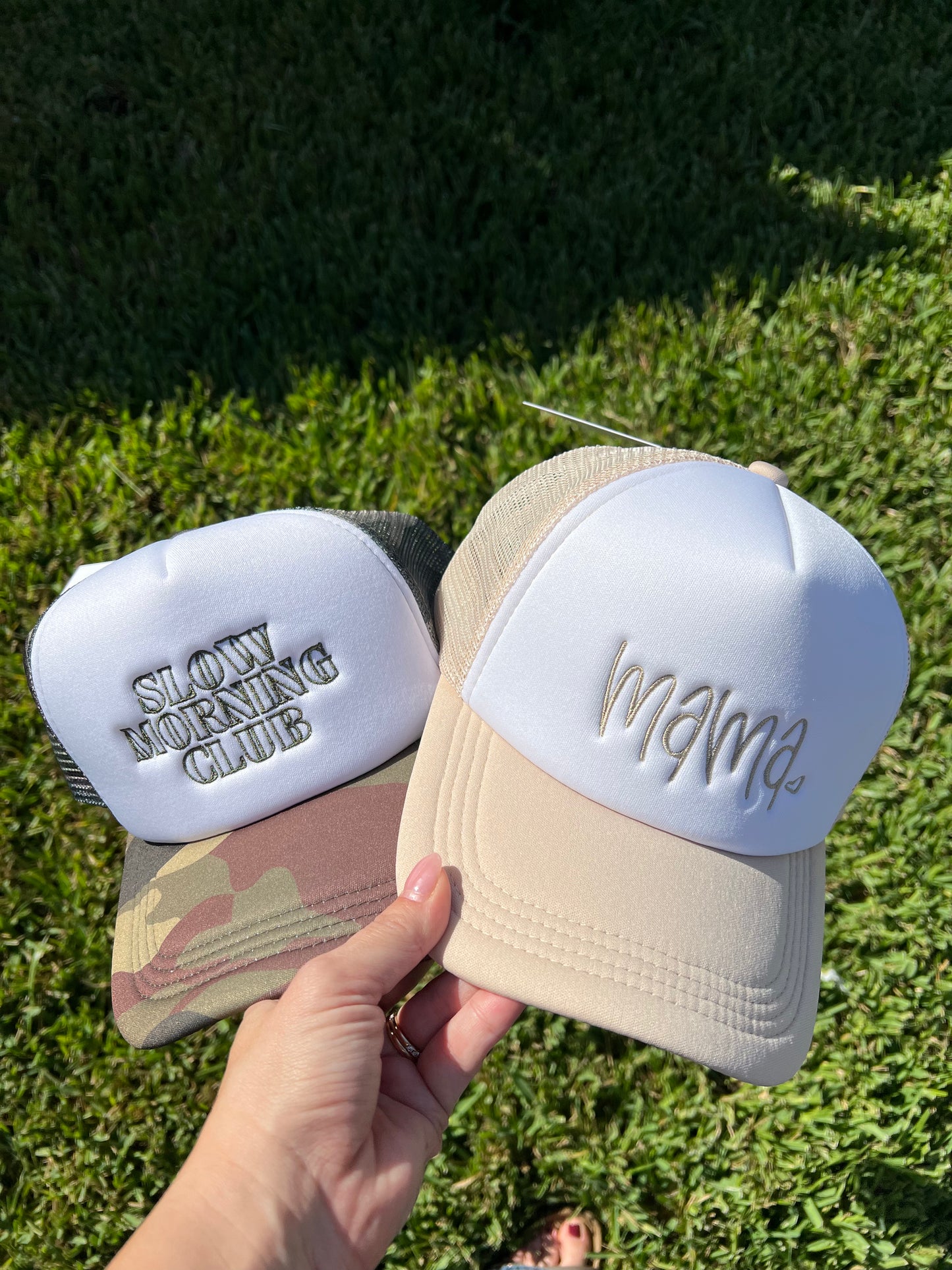 Two hats, one with 'Slowly Morning Club' and the other with 'mama', held against a grassy background.