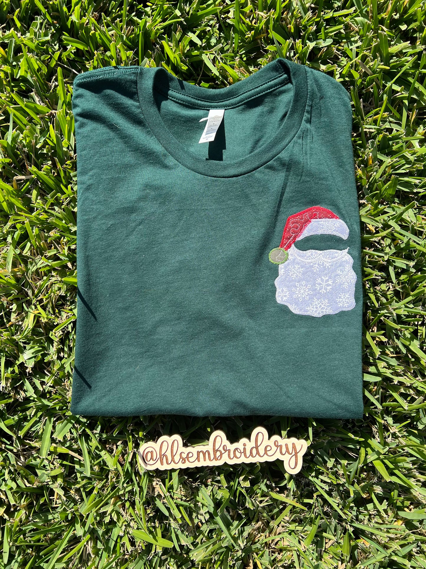 Green t-shirt with a Santa Claus design on grass, featuring the brand 'hlisembroidery'.