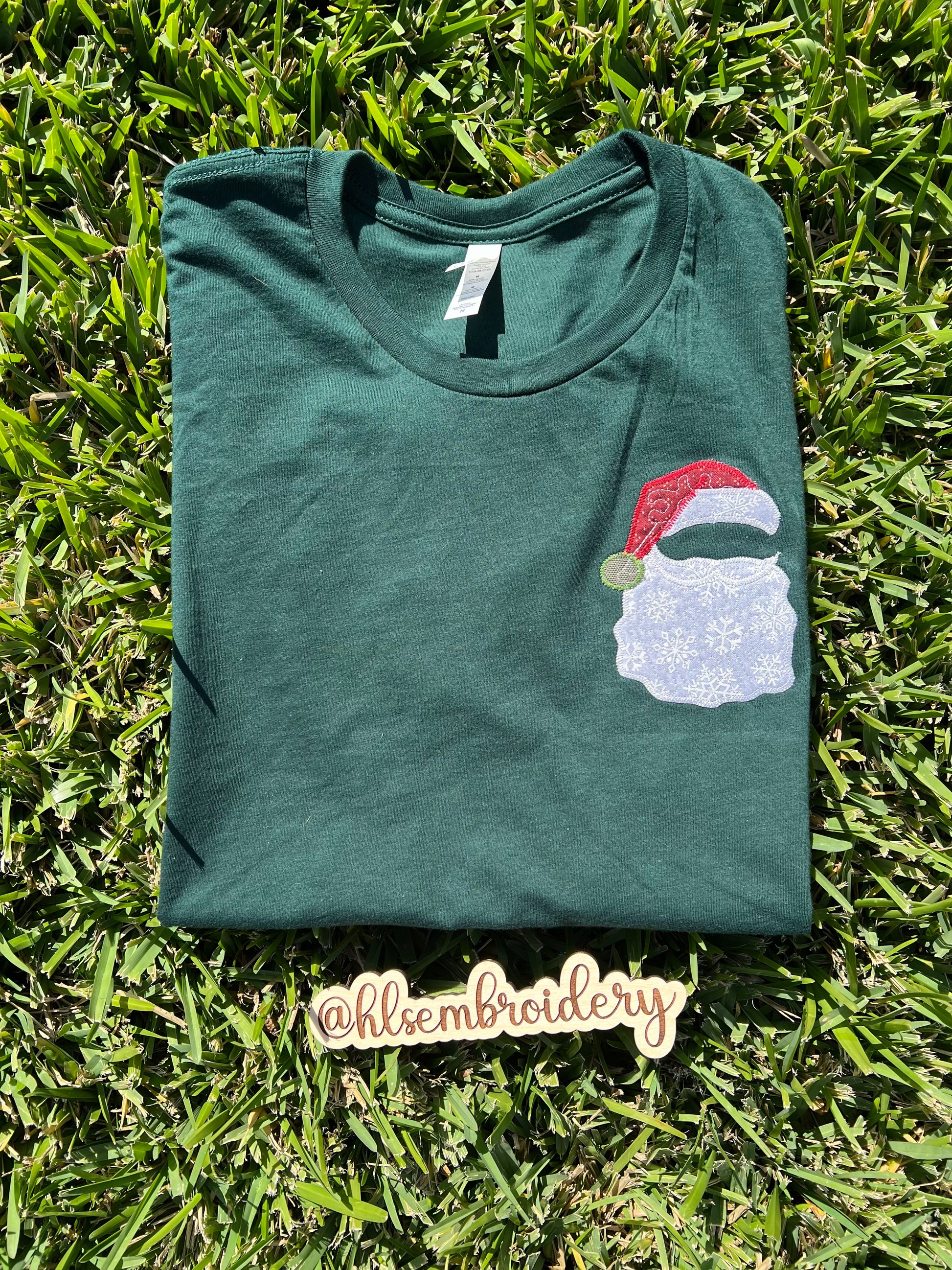 Green t-shirt with a Santa Claus design on grass, featuring the brand 'hlisembroidery'.