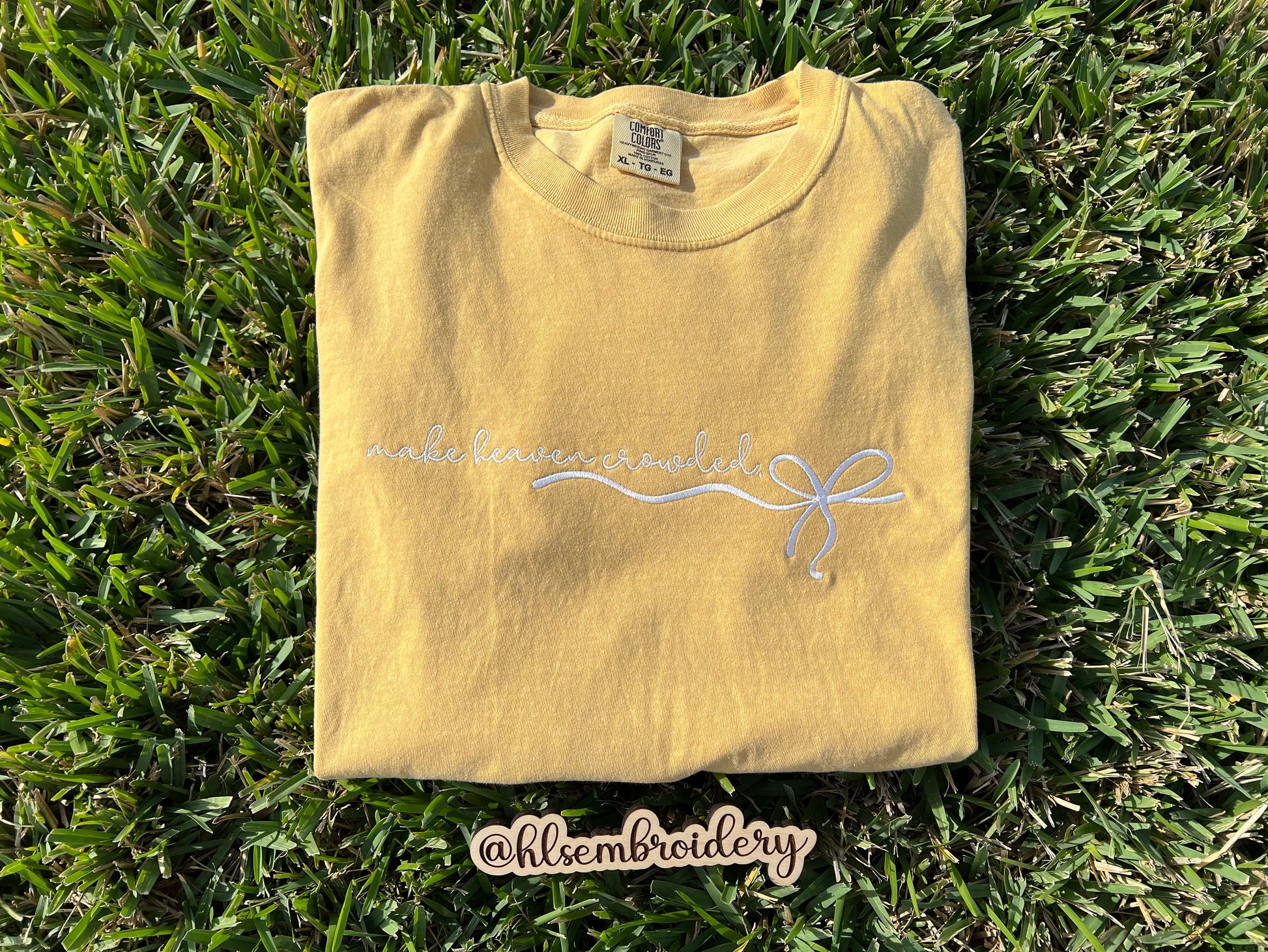Folded yellow t-shirt with embroidery on grass, featuring a brand name.