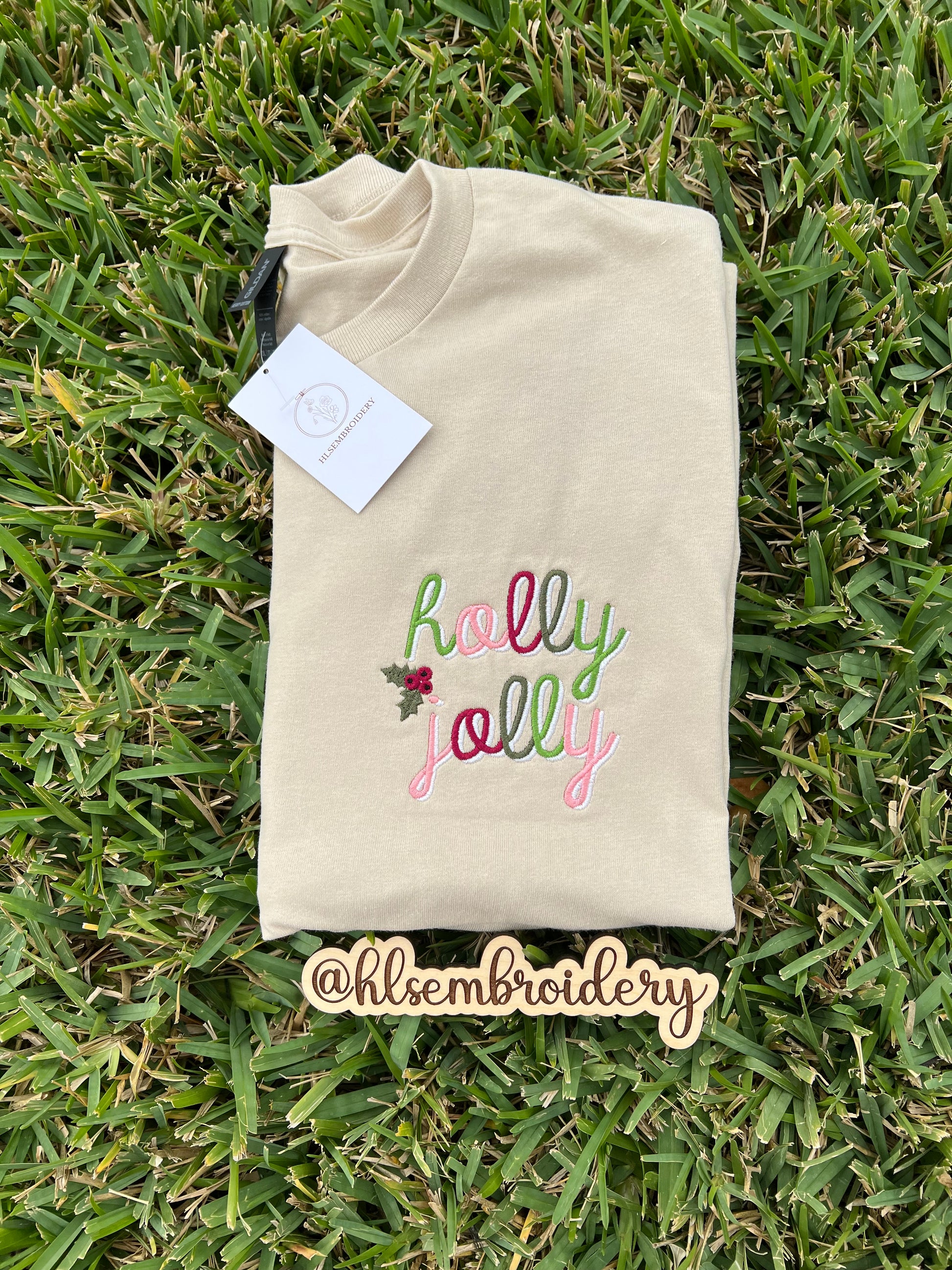 Folded sand color shirt with 'holly jolly' embroidery on grass, featuring the brand '@hlsembroidery'.