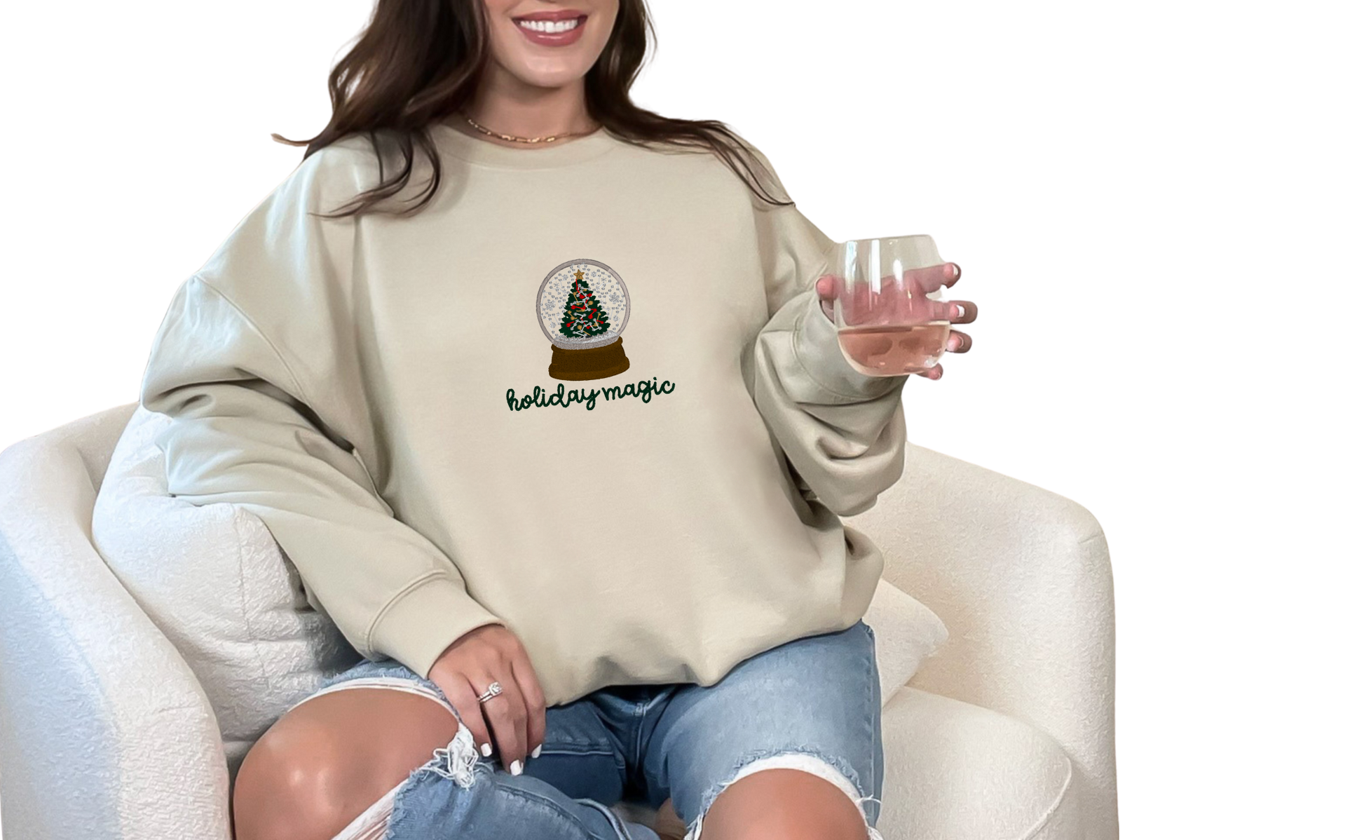 Woman wearing a sand color sweatshirt with 'holiday magic' design, sitting on a white chair.