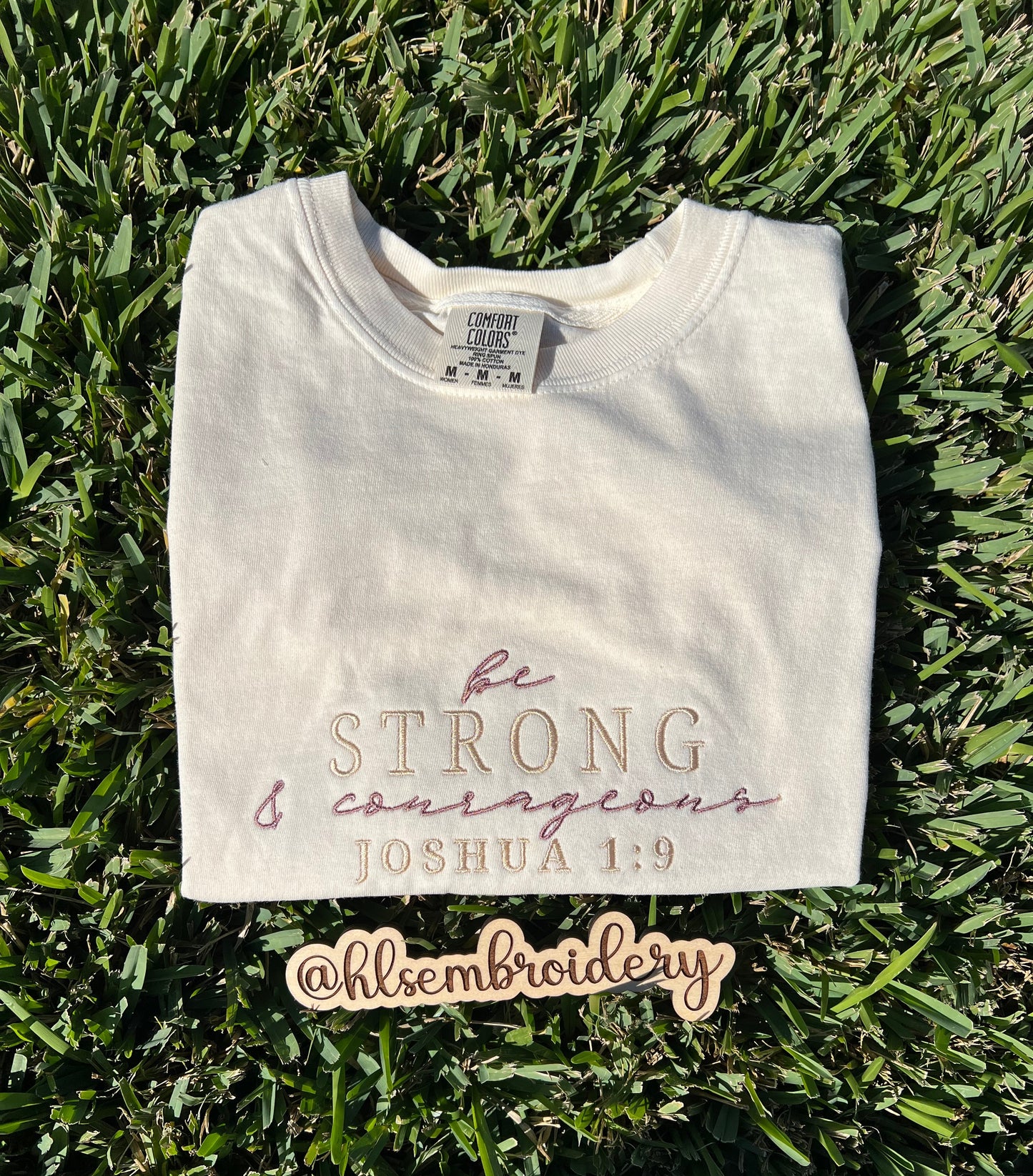 Folded Ivory t-shirt with 'be strong and courageous' text on grass