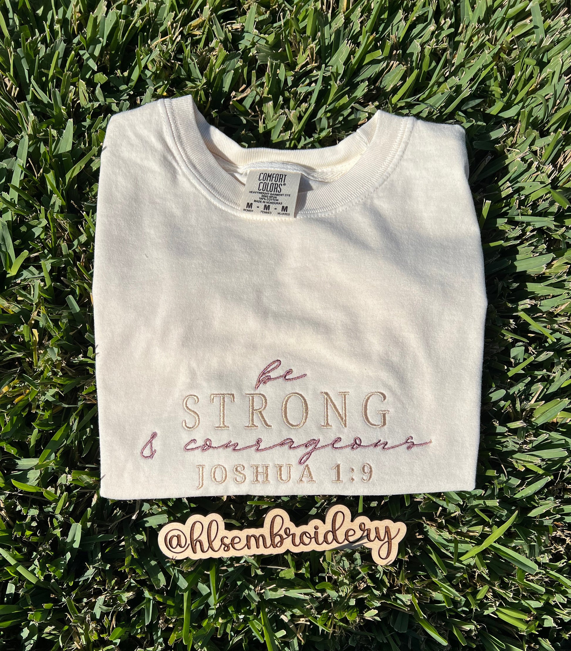 Folded Ivory t-shirt with 'be strong and courageous' text on grass