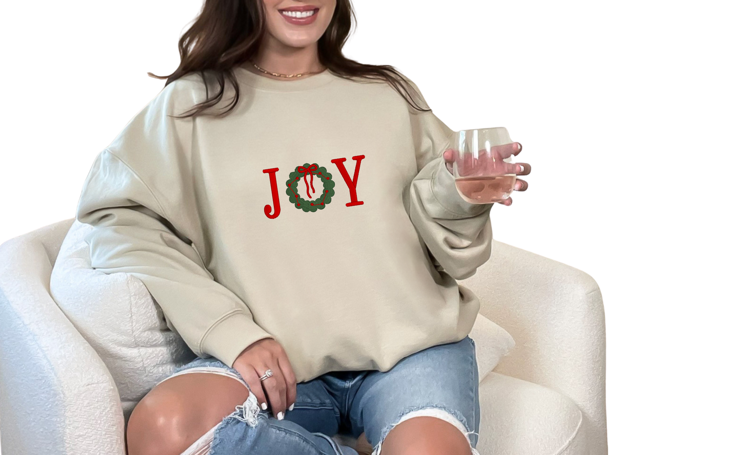 Person wearing a sand color sweatshirt with 'JOY' and a wreath design in the center, sitting on a white chair.
