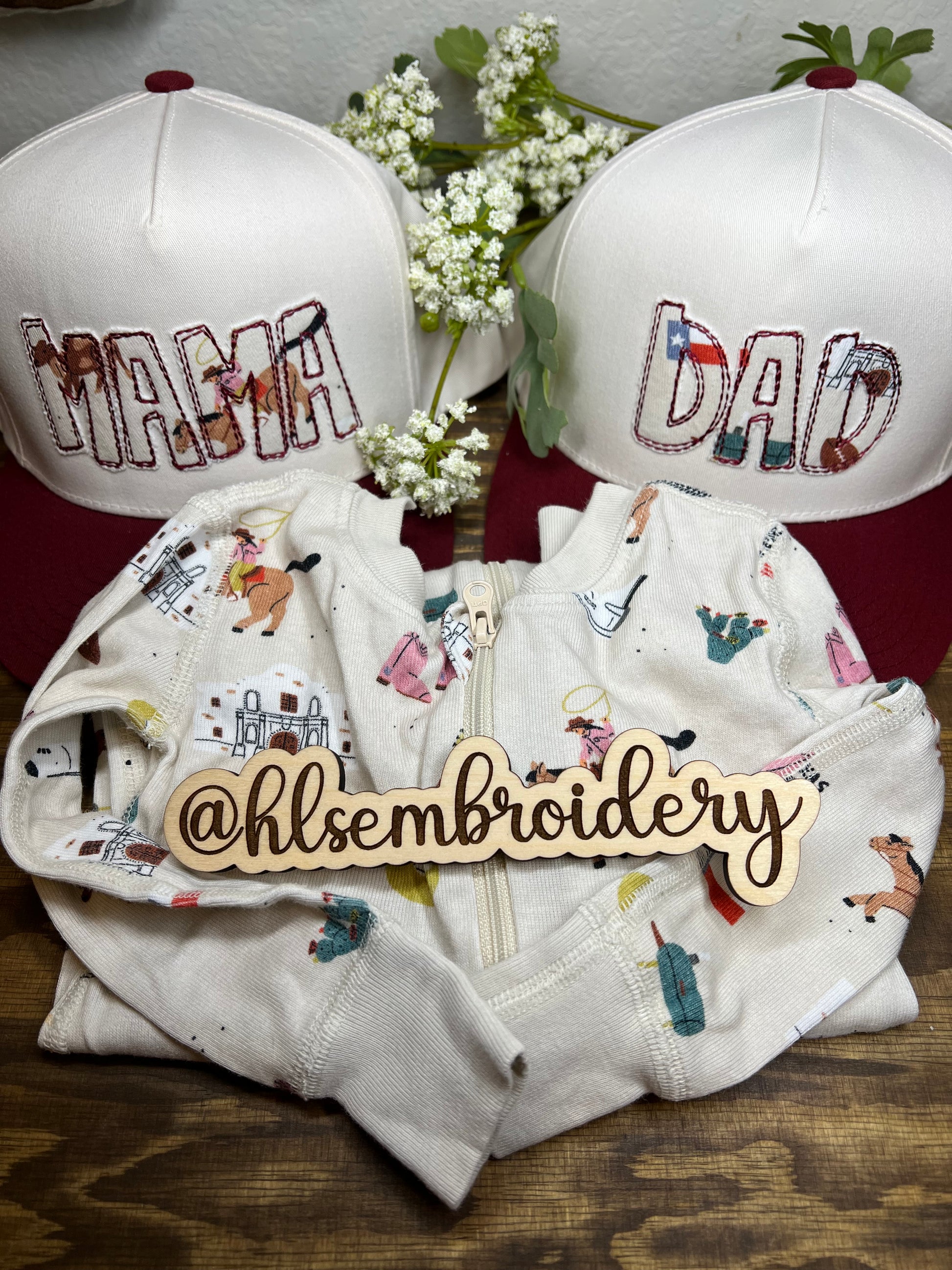 Embroidered hats with 'DAD' and 'MAMA' on a wooden surface with floral decorations.