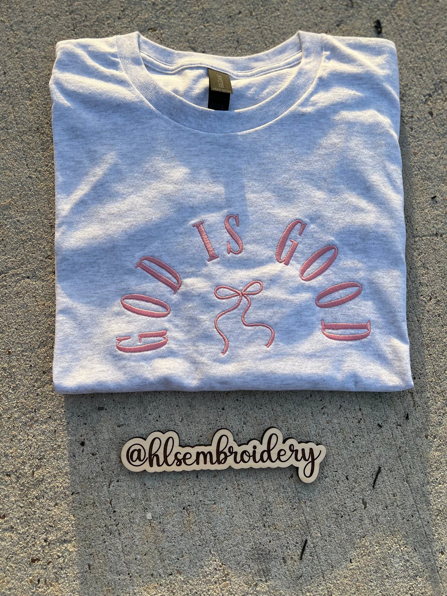 Folded white t-shirt with 'GOD IS GOOD' text and embroidered design on a concrete surface, with 'hlsembrdery' logo below.