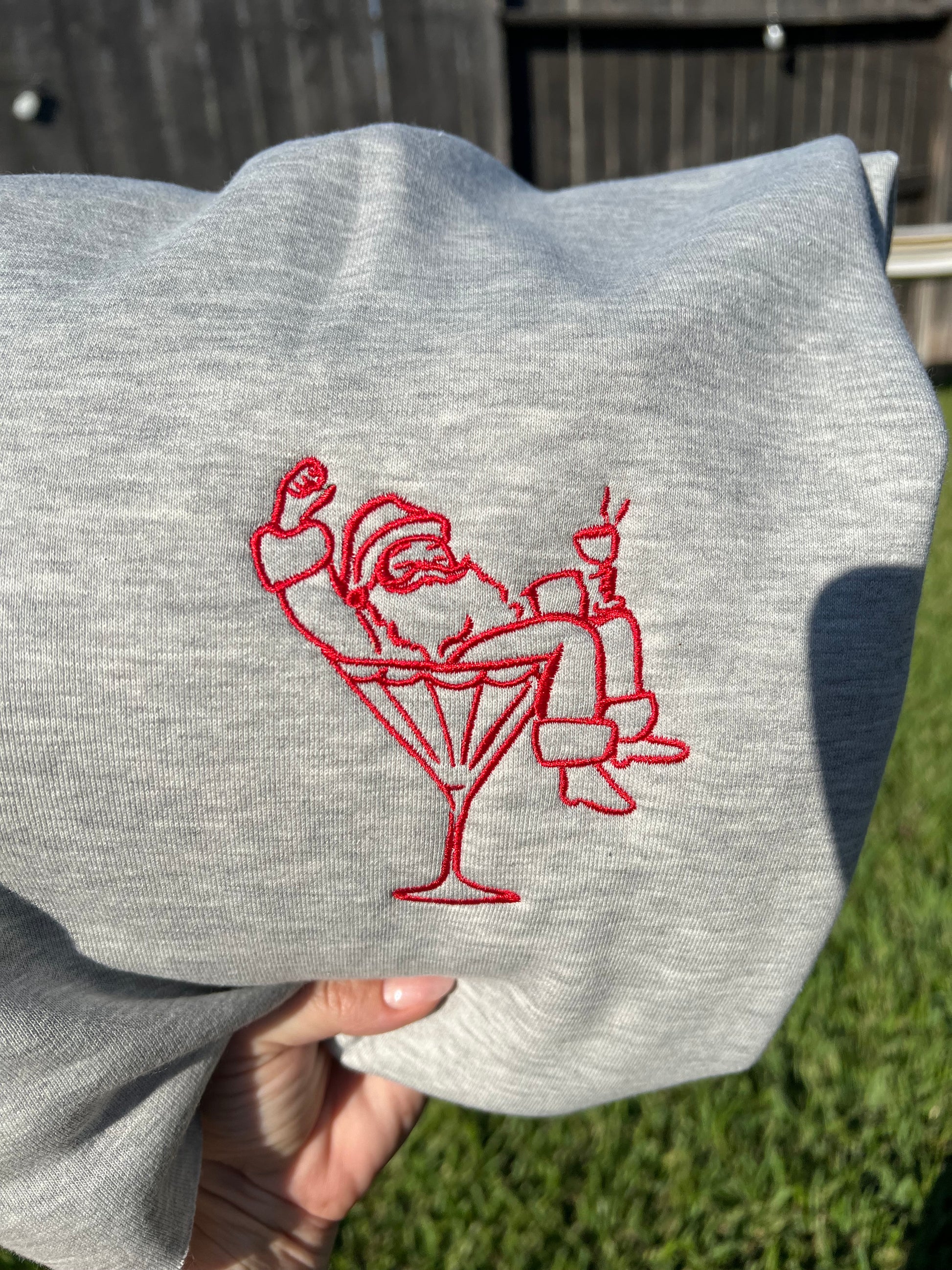 Gray sweatshirt with red embroidered design of santa lounging in a martini glass, held against a blurred outdoor background.