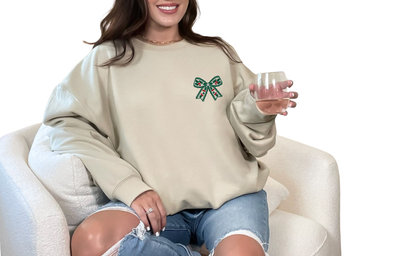Woman wearing a sand color sweatshirt with a green bow design, sitting on a white chair.