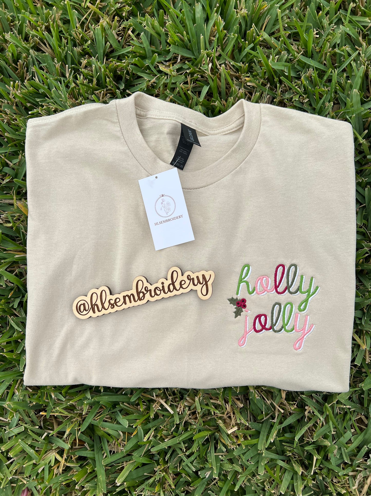 Folded sand color shirt with 'holly jolly' embroidery on a concrete surface, featuring the brand '@hlsembroidery'.