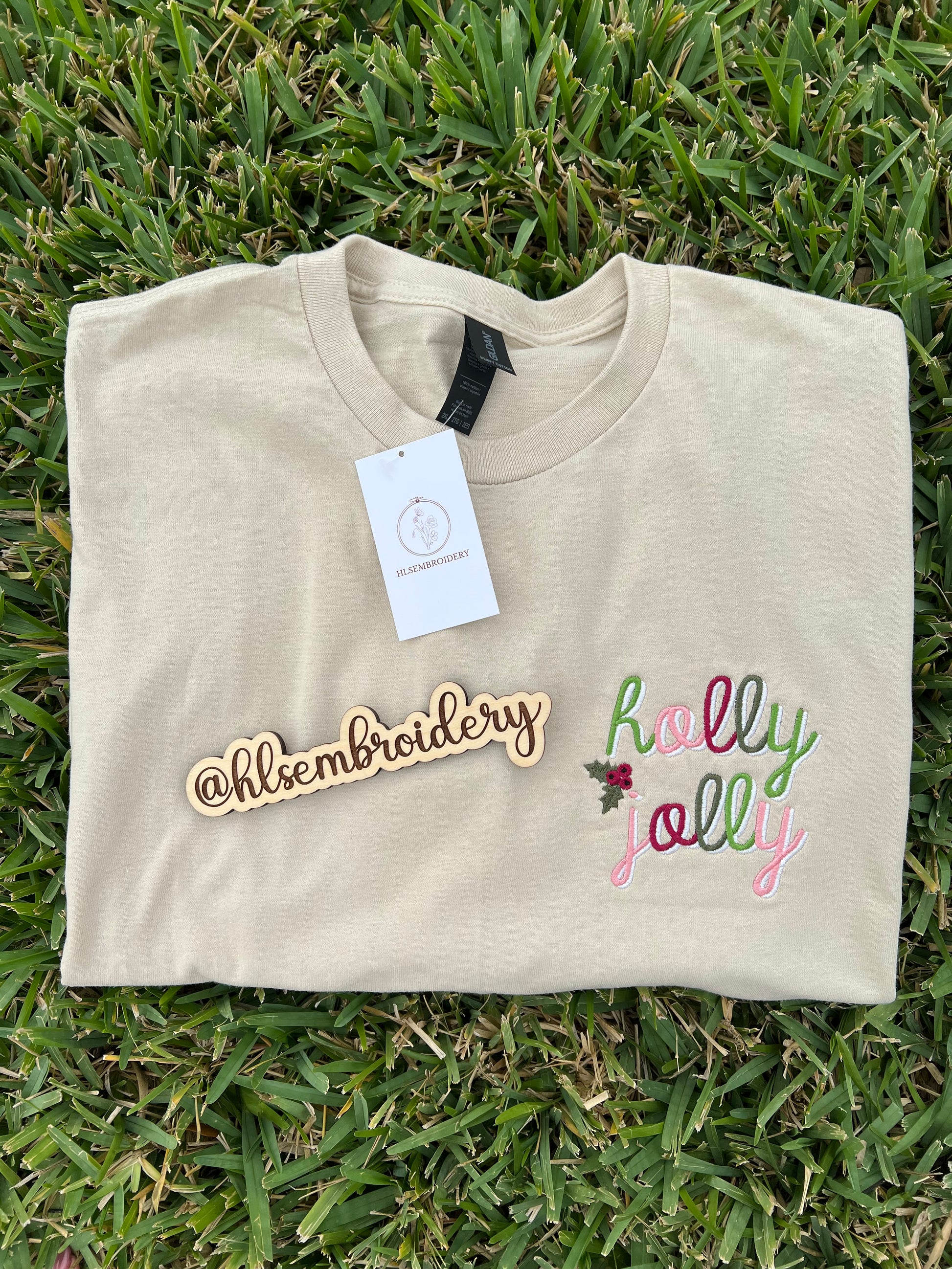 Folded sand color shirt with 'holly jolly' embroidery on a concrete surface, featuring the brand '@hlsembroidery'.