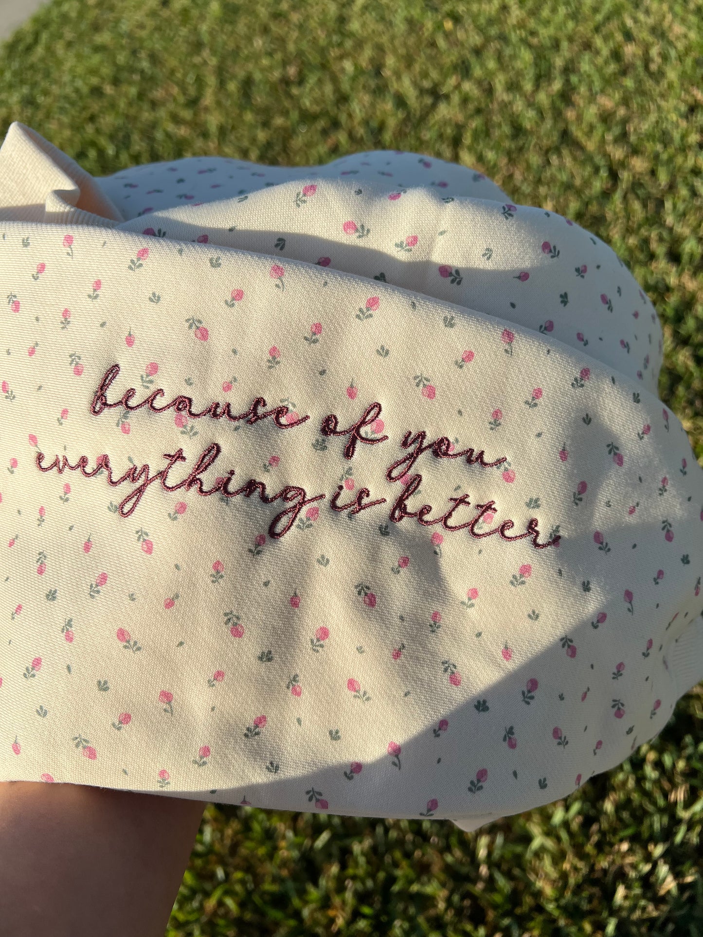 Floral-patterned fabric with embroidered text "because of you everything is better" held against a grassy background