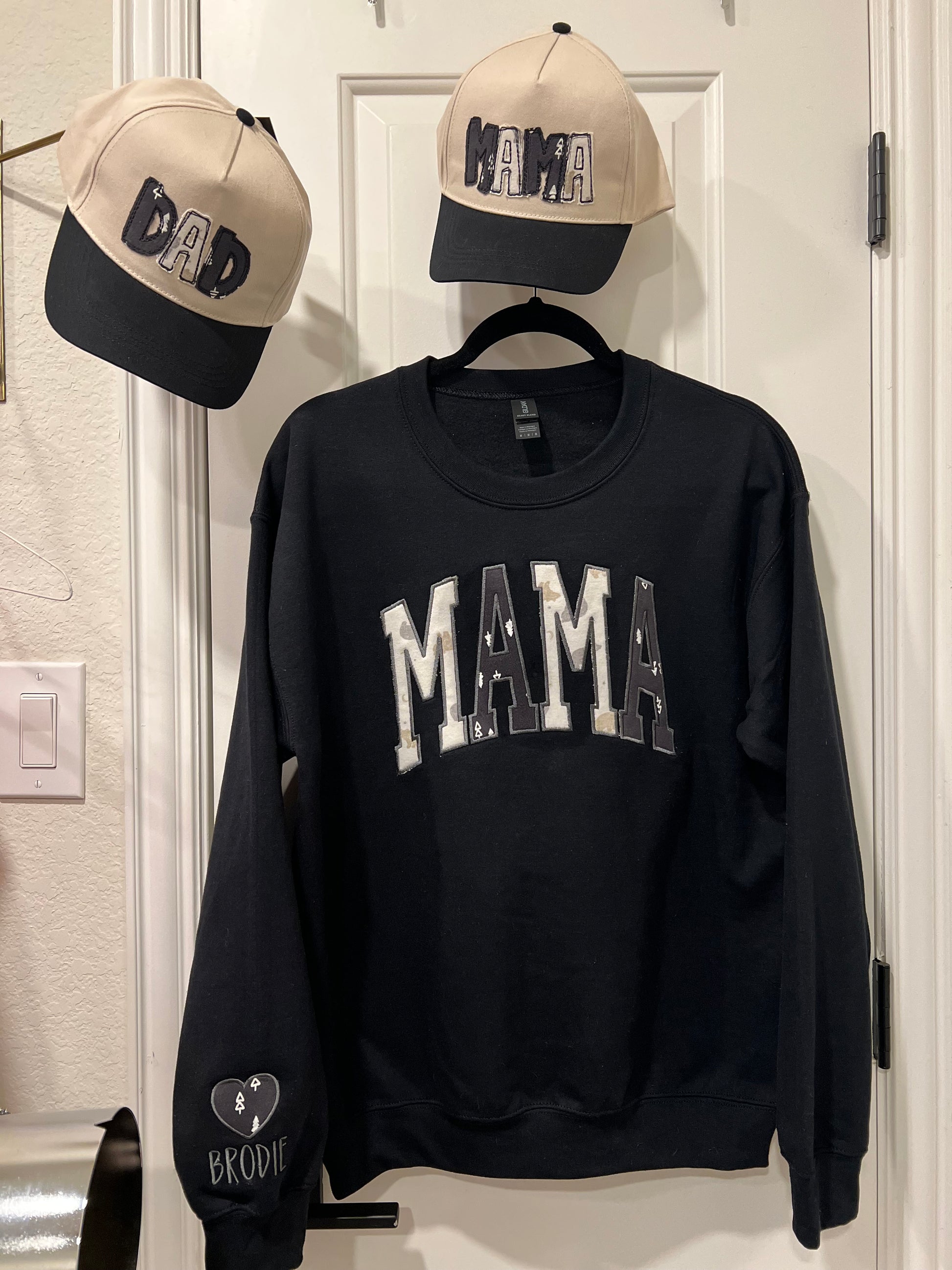 Black sweatshirt with 'MAMA' text, beige cap with black text, and black cap with white text on a light-colored wall.