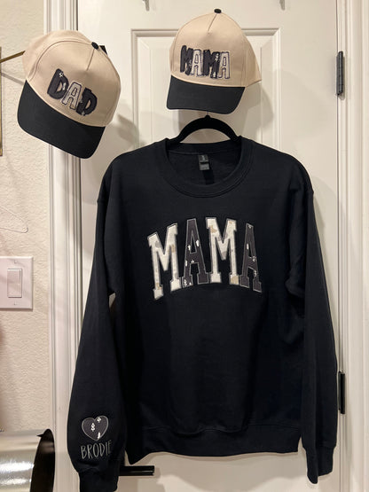 Black sweatshirt with 'MAMA' text, beige cap with black text, and black cap with white text on a light-colored wall.
