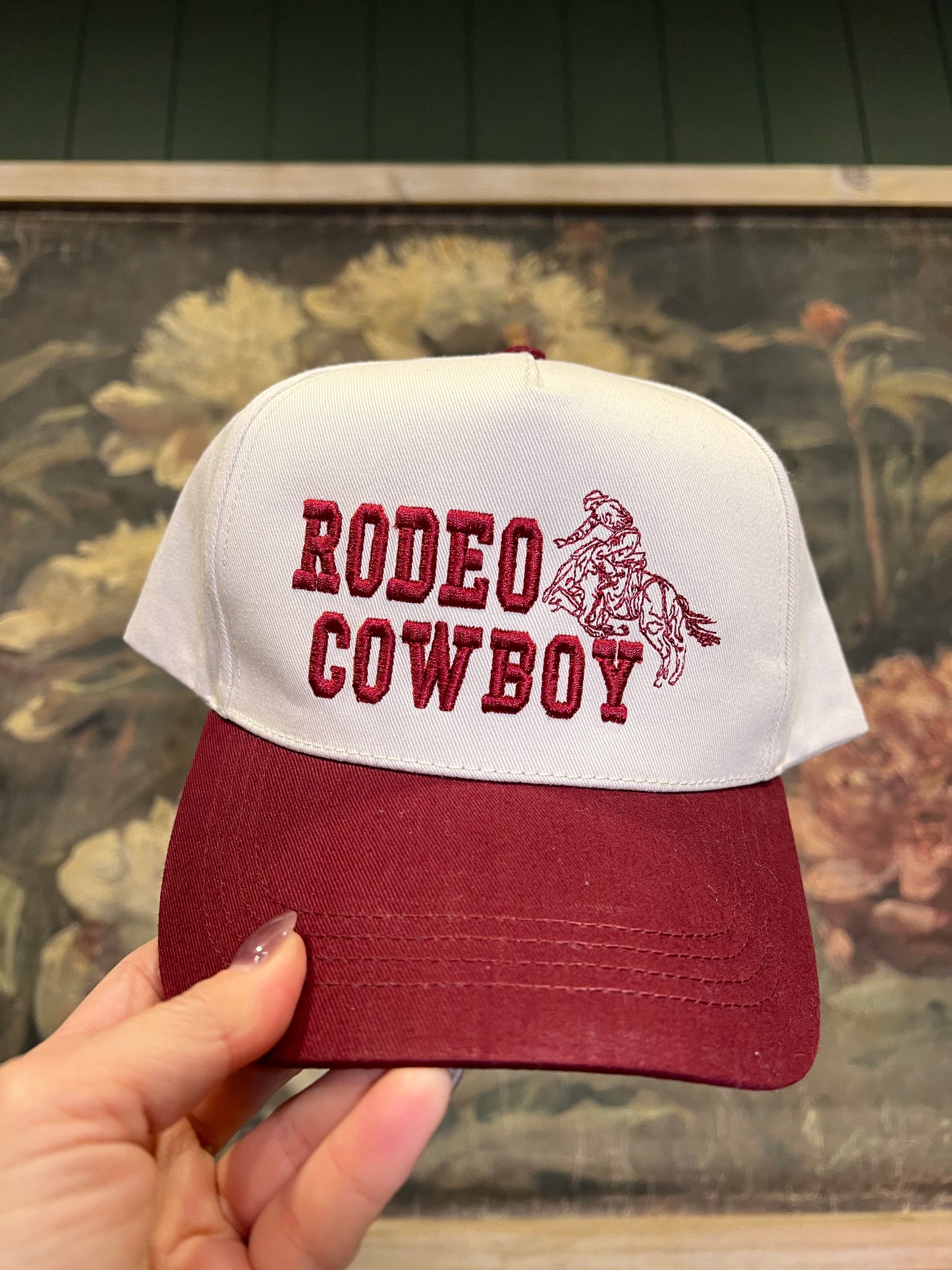 Cap with 'Rodeo Cowboy' text and logo held by a hand against a floral background