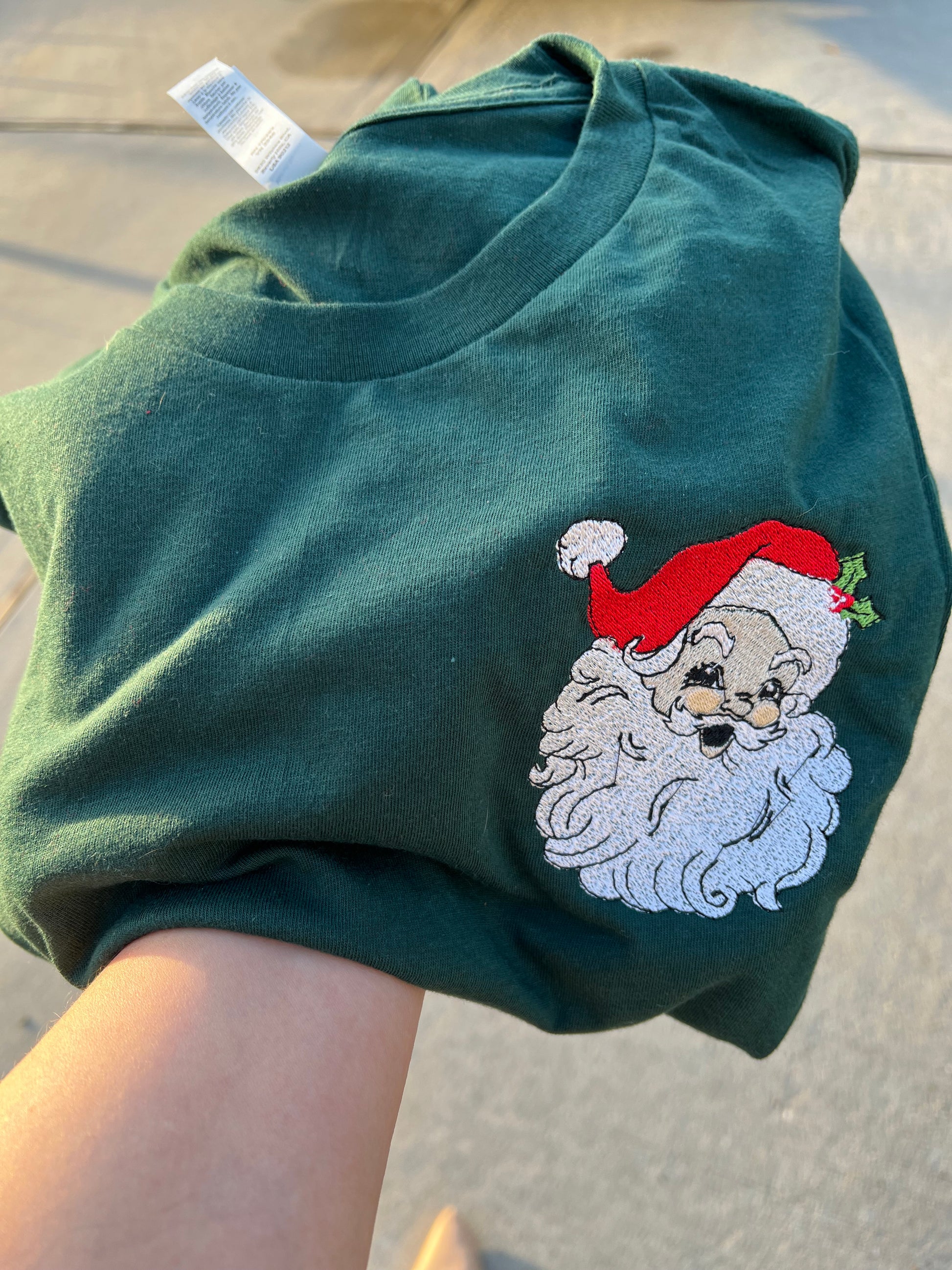 Folded green t-shirt with embroidered Santa Claus design on a concrete surface, featuring the brand '@hlsembrdery'.