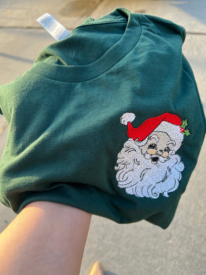 Folded green t-shirt with embroidered Santa Claus design on a concrete surface, featuring the brand '@hlsembrdery'.