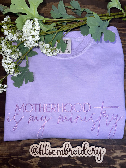 Purple garment with 'Motherhood is my ministry' embroidery on a wooden surface with greenery.