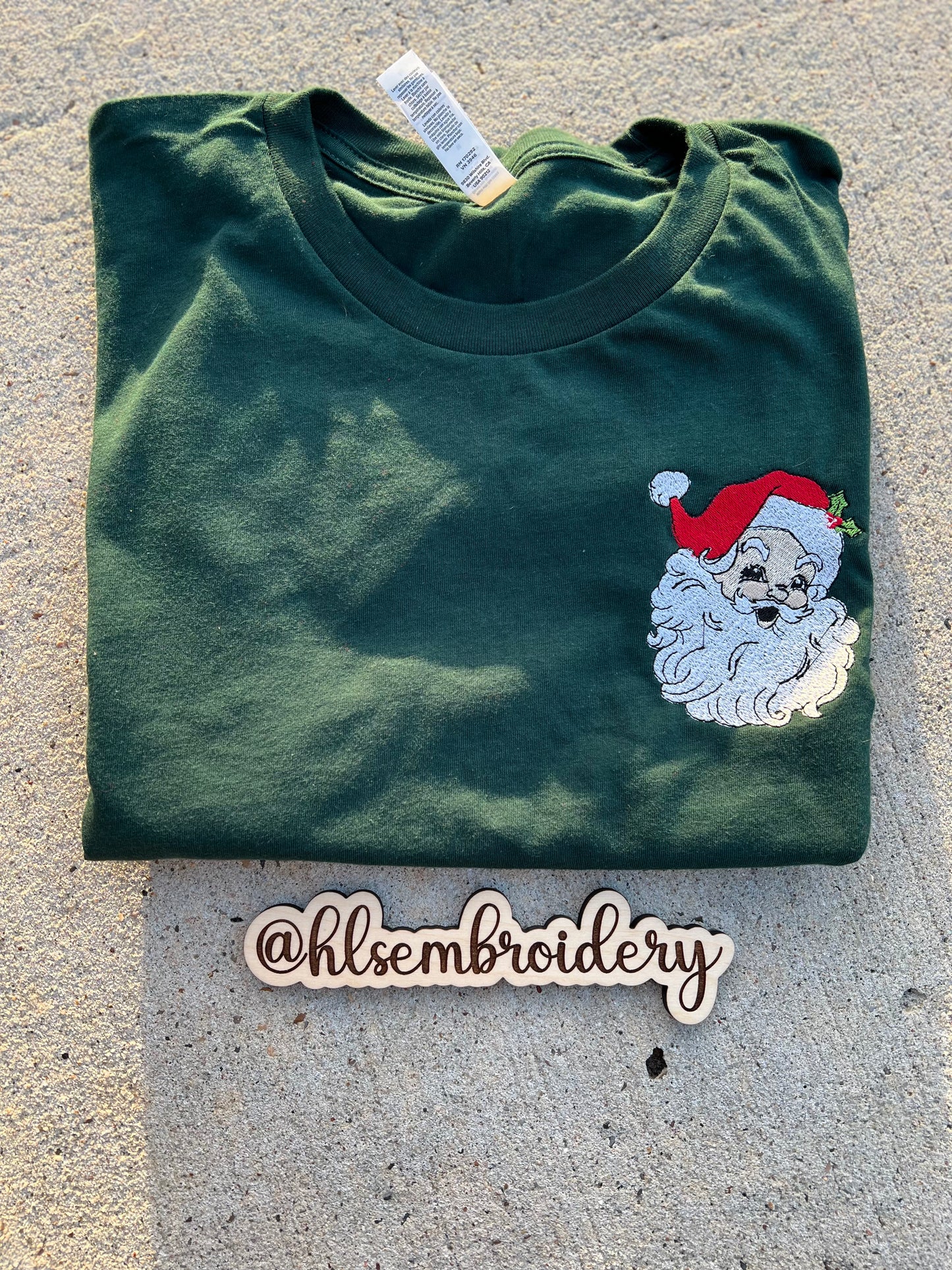 Folded green t-shirt with embroidered Santa Claus design on a concrete surface, featuring the brand '@hlsembrdery'.
