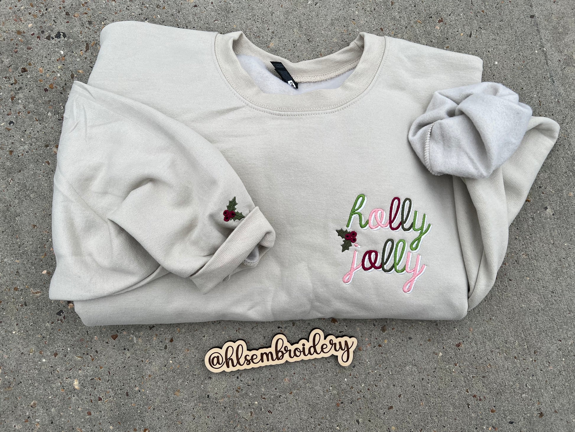 Folded sand color sweatshirt with 'holly jolly' embroidery on a concrete surface, featuring the brand '@hlsembroidery'.