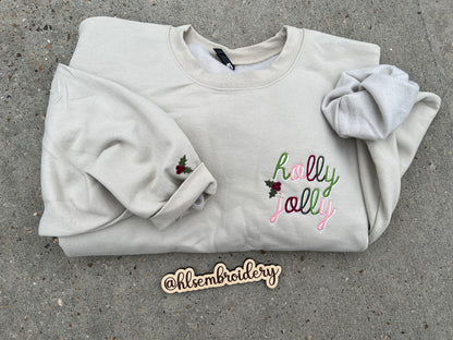 Folded sand color sweatshirt with 'holly jolly' embroidery on a concrete surface, featuring the brand '@hlsembroidery'.