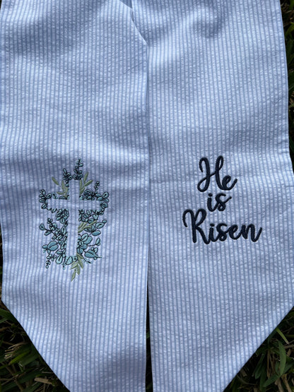 Embroidered wreath sash with cross and 'He is Risen' text on a textured fabric background