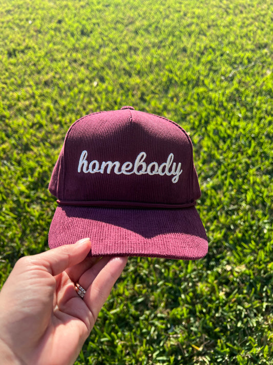 Maroon corduroy cap with 'homebody' text embroidery held against a blue sky