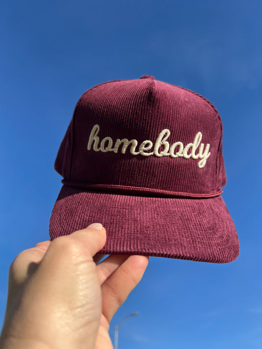 Maroon corduroy cap with 'homebody' text embroidery held against a blue sky