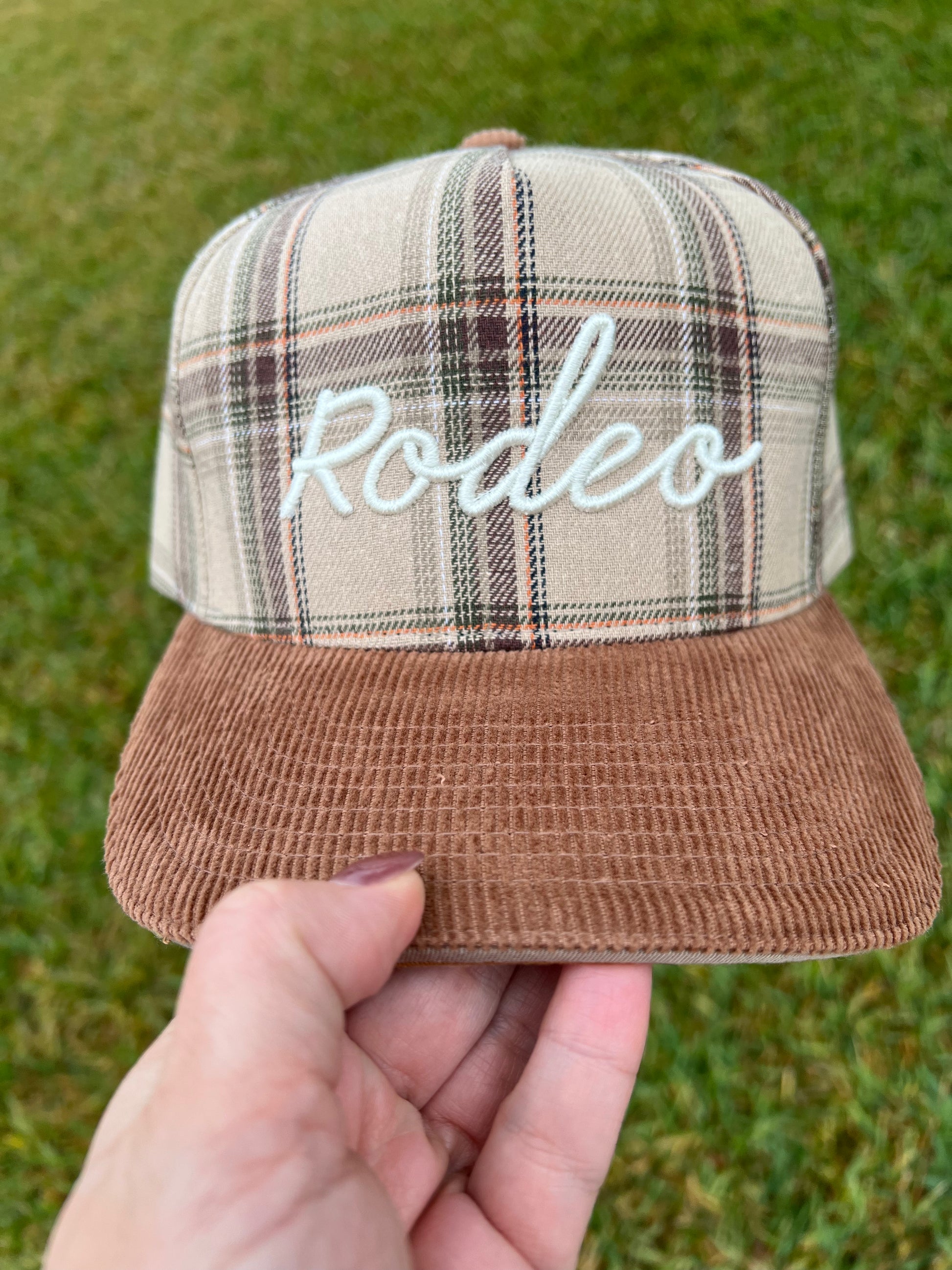 Plaid cap with 'Rodeo' embroidery held by a hand on grass