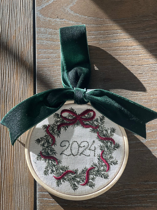 Embroidery hoop with a wreath design and '2024' text, tied with a green ribbon on a wooden surface.