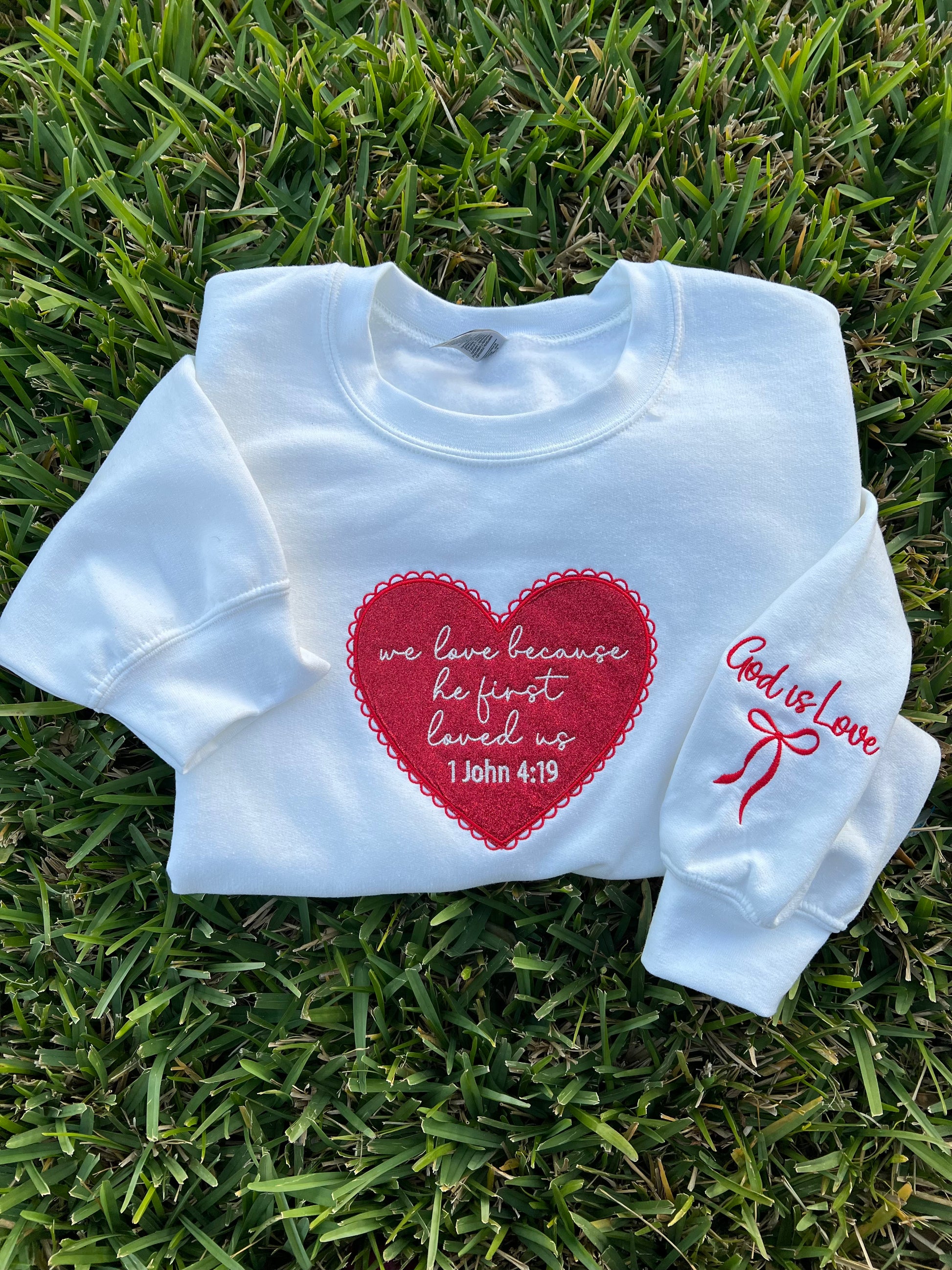 White sweatshirt with red heart design and text embroidery "we love because he first loved us 1 John 4:19" and  "God is Love" text and bow embroidery on the sleeve. 