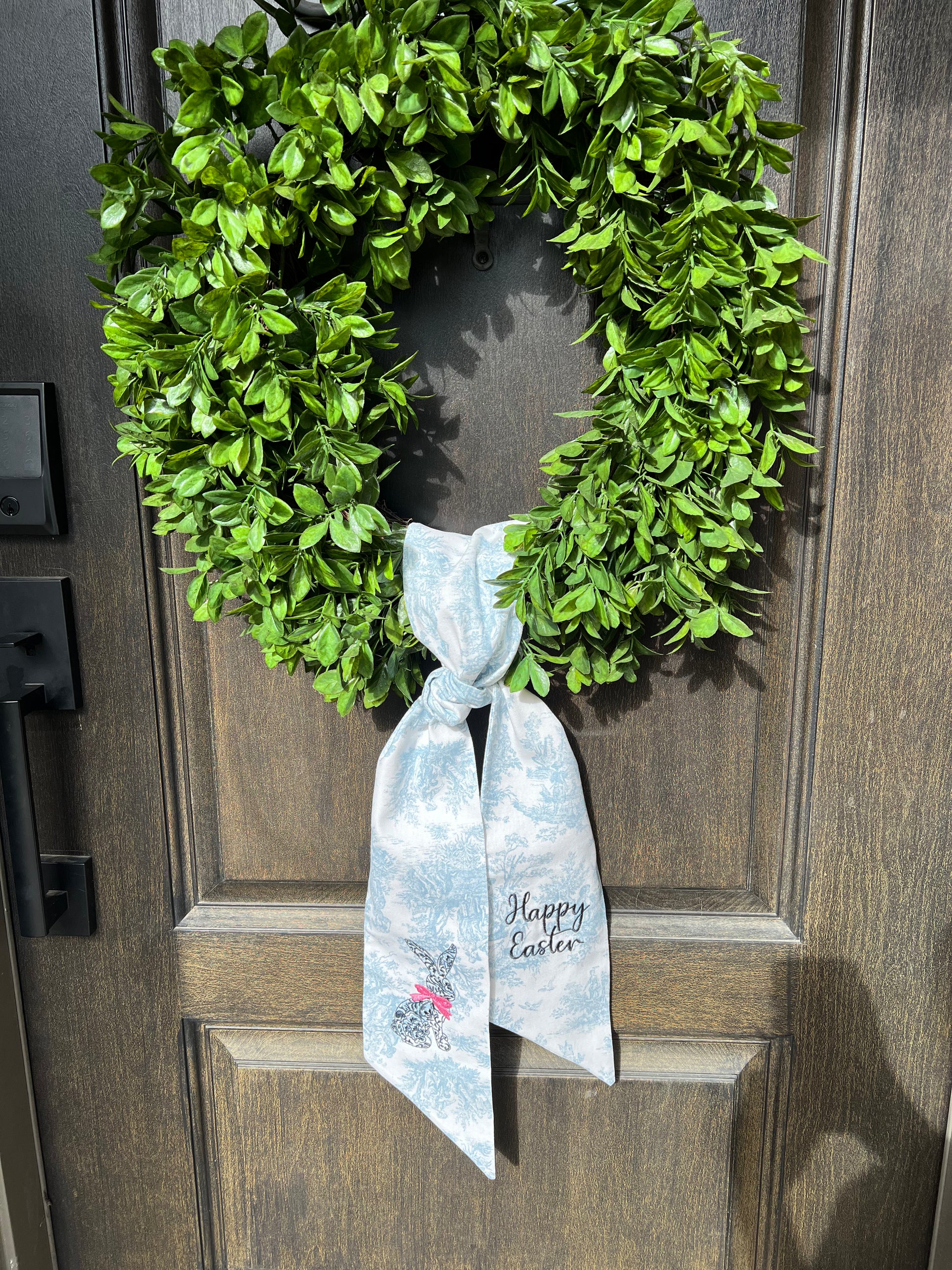 Blue toile wreath sash with a rabbit design and 'Happy Easter' text shown hanging on a wreath tied and hanging in the front door