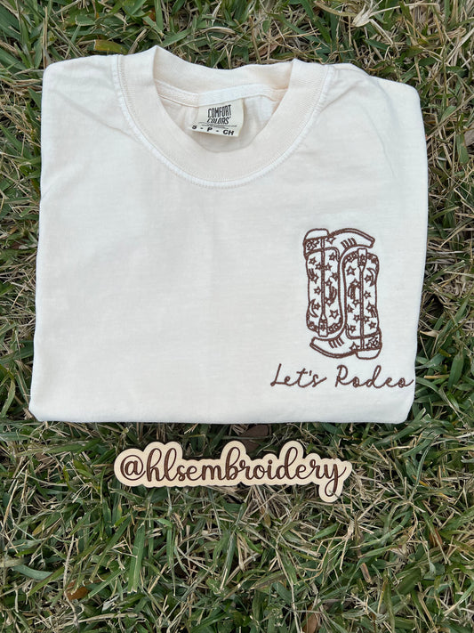 Ivory shirt with lets rodeo wording and boots embroidered on it 