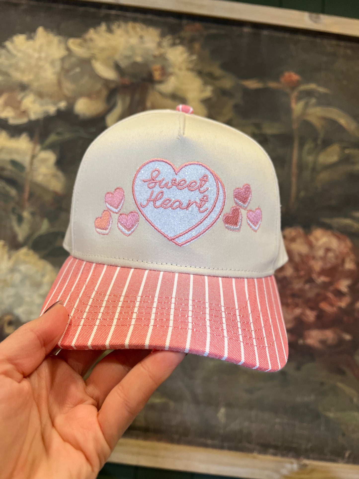 Pink and white cap with 'sweet heart' embroidery held in front of a floral background