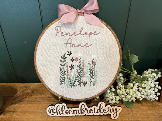 Embroidery hoop with floral design and embroidered name, featuring a pink bow and white flowers on a wooden surface.