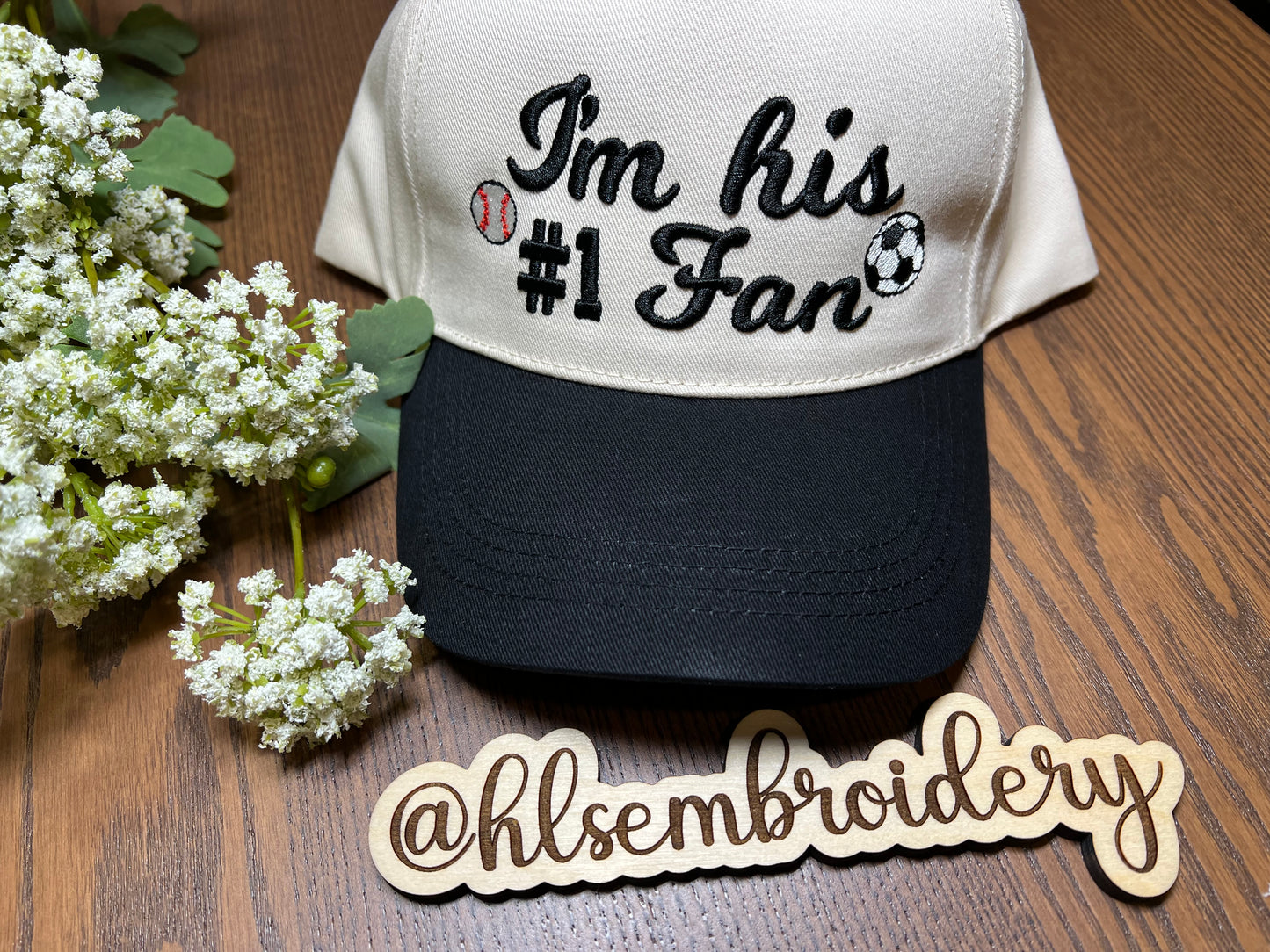 Cap with 'I'm his #1 Fan' text on a wooden surface with flowers, featuring @hlsembroidery logo.