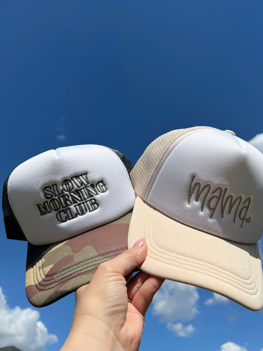 Two caps, one with 'Slowly Morning Club' and the other with 'mama', held against a blue sky.