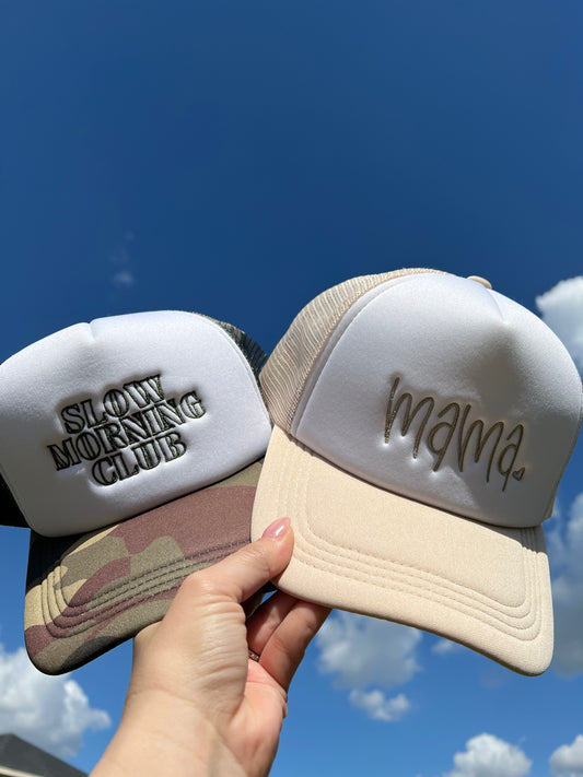 Two hats, one with 'Slovy Morning Club' and the other with 'mama', held against a blue sky.