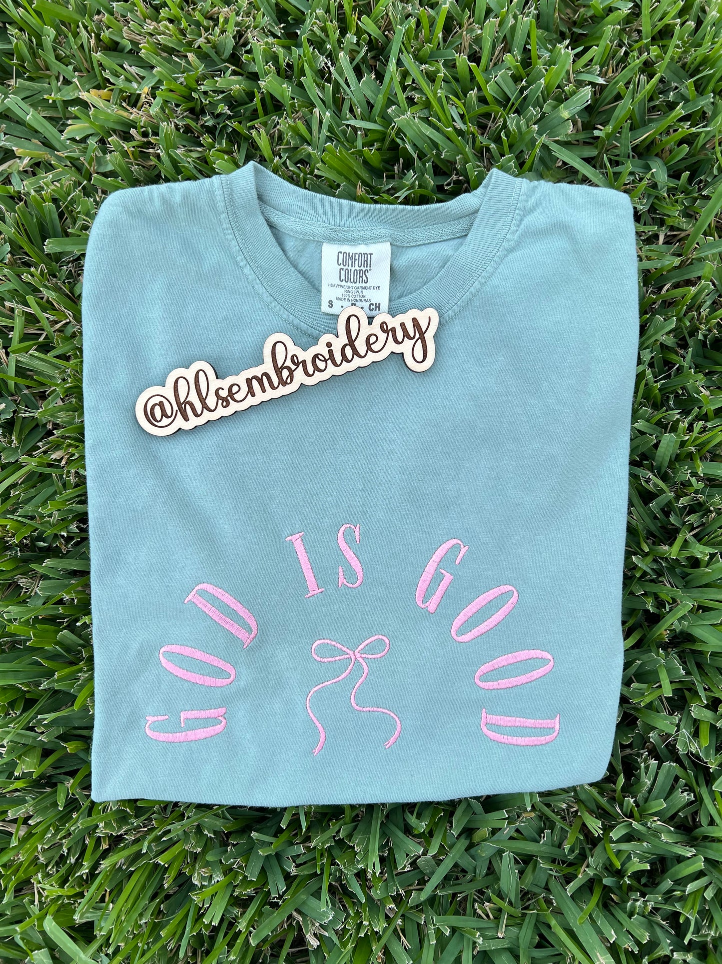 Light blue t-shirt with 'GOD IS GOOD' text and bow design on grass