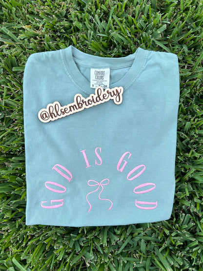 Light blue t-shirt with 'GOD IS GOOD' text and bow design on grass