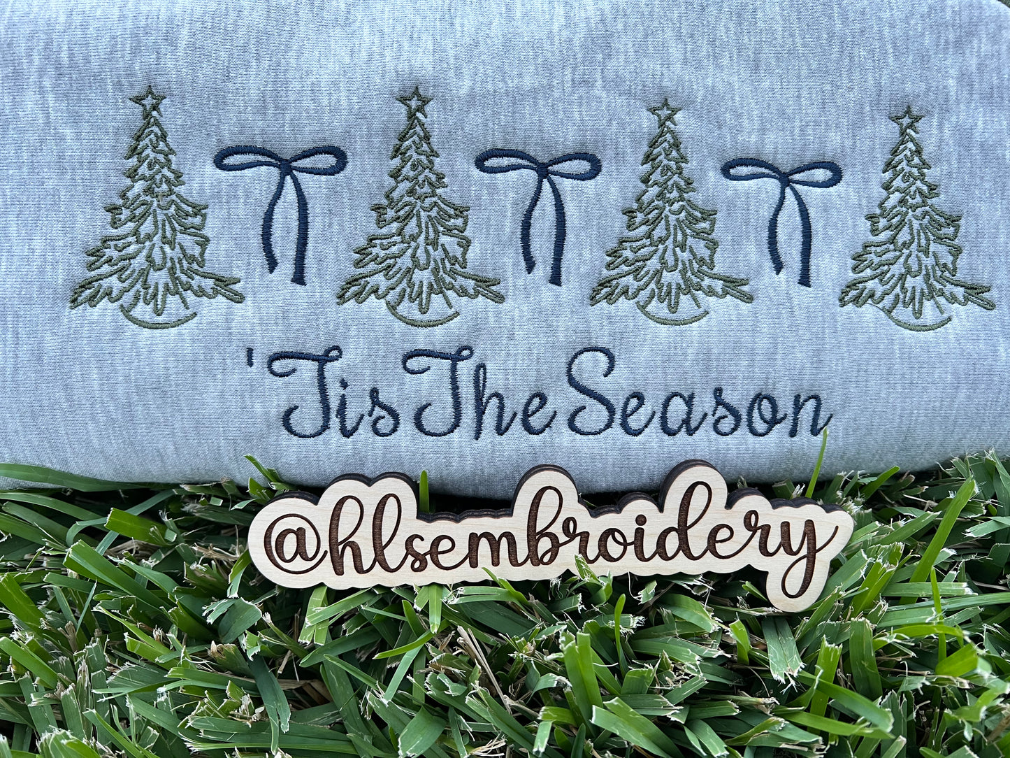 Embroidered design with trees, bows, and 'Tis The Season text on fabric, with grass at the bottom.