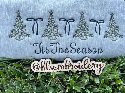 Embroidered design with trees, bows, and 'Tis The Season text on fabric, with grass at the bottom.