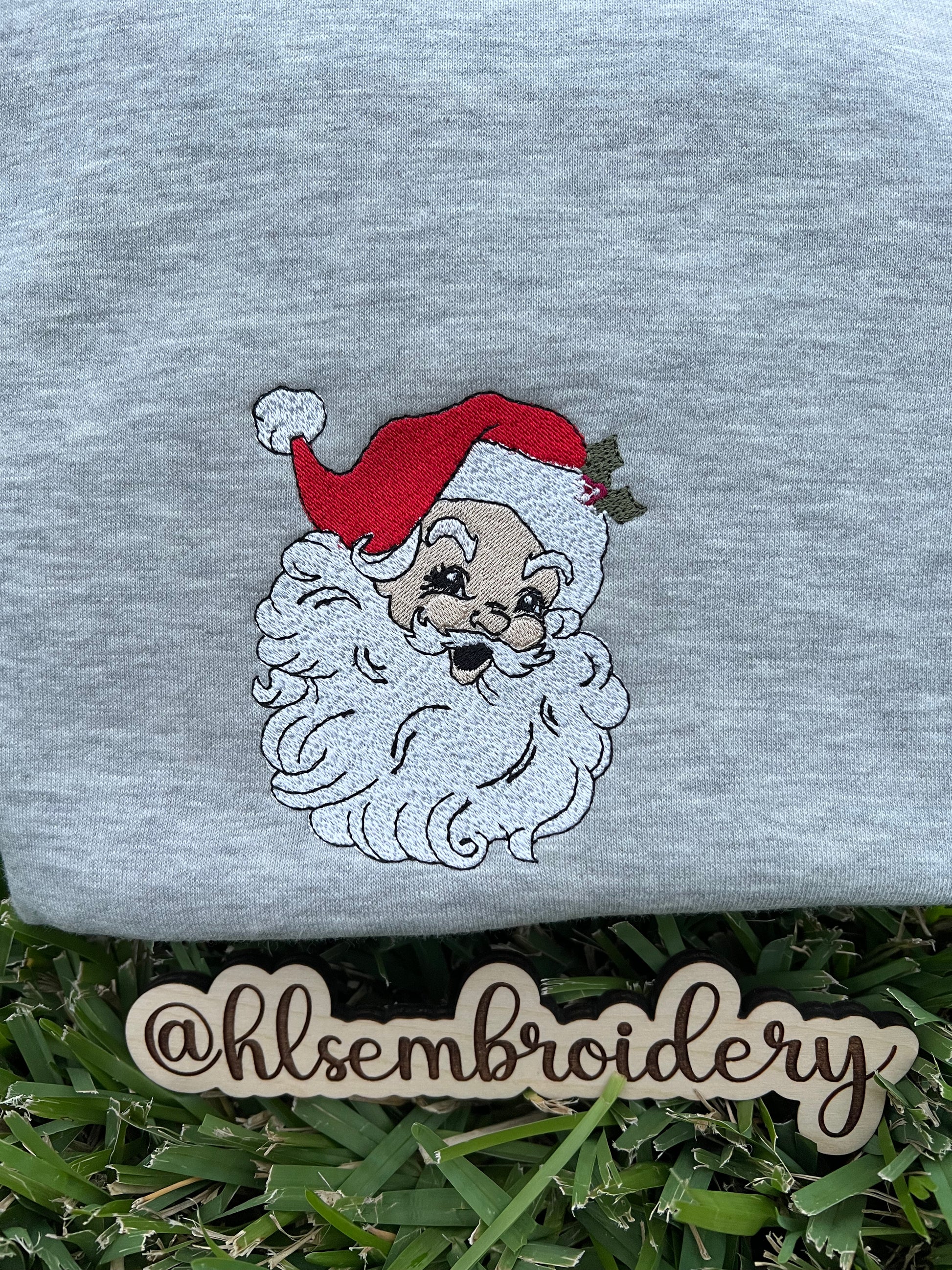 Gray sweatshirt with embroidered Santa Claus face on grass, featuring the brand 'hlsembrdery'.