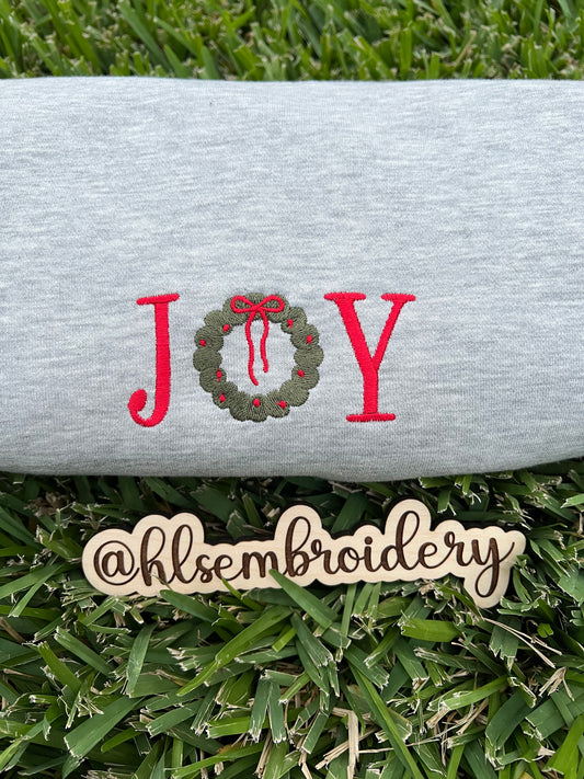 Embroidered word 'JOY' with a wreath design on a piece of fabric, placed on grass with a wooden sign displaying '@hlsembroidery'.