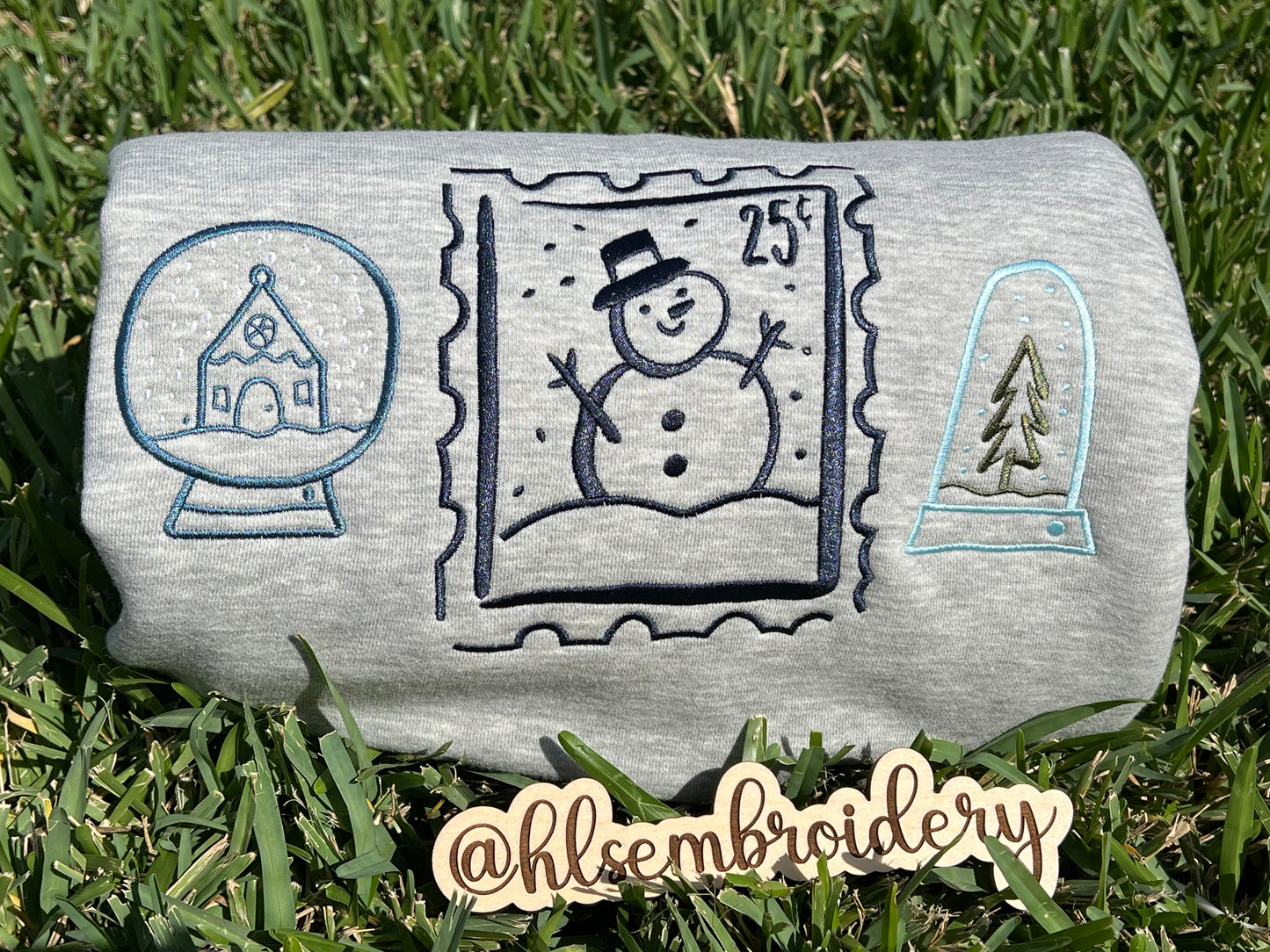 Embroidered designs of a snowman, snow globe, and pine tree on grass.