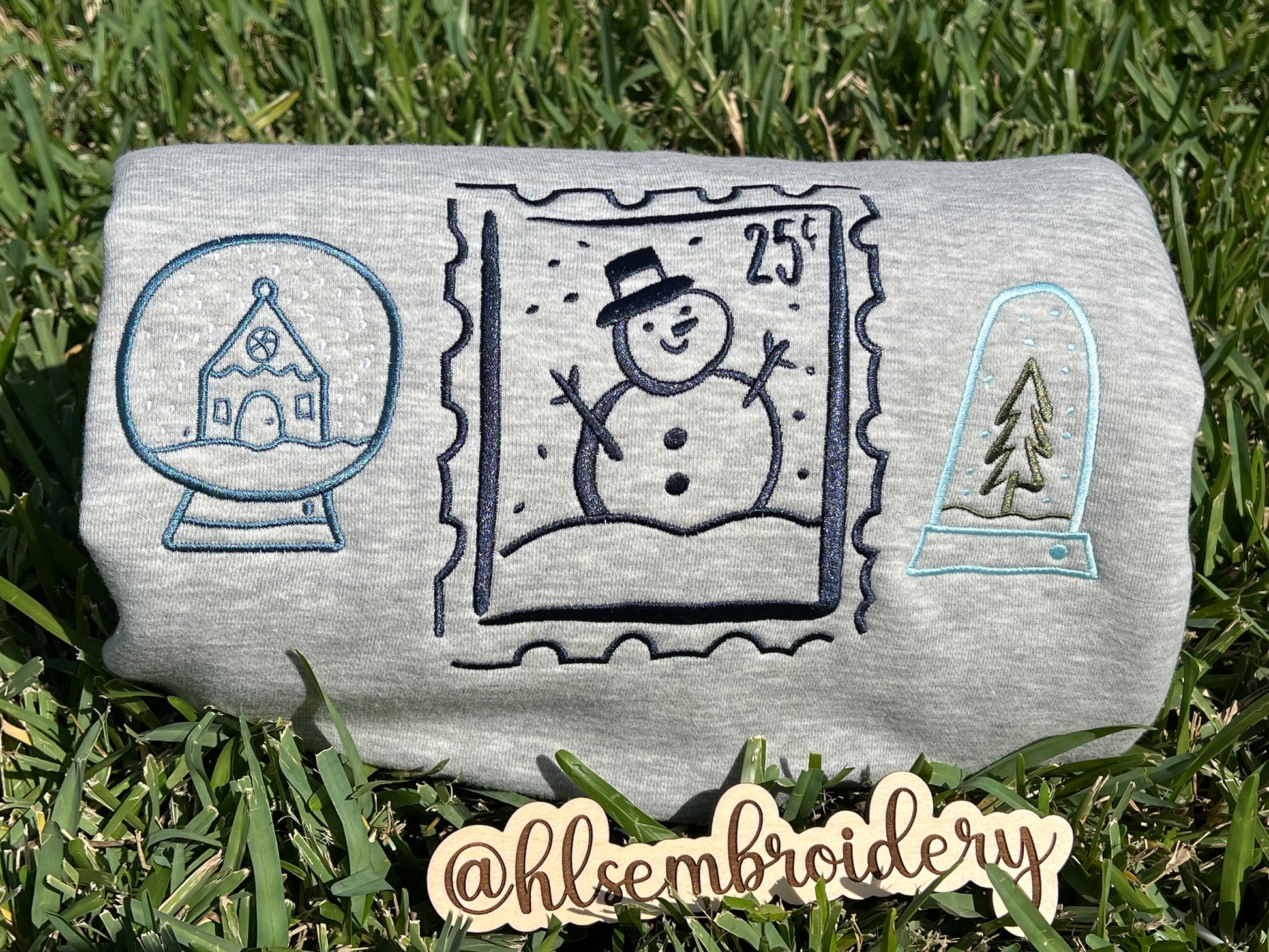 Embroidered designs of a snowman, snow globe, and pine tree on grass.