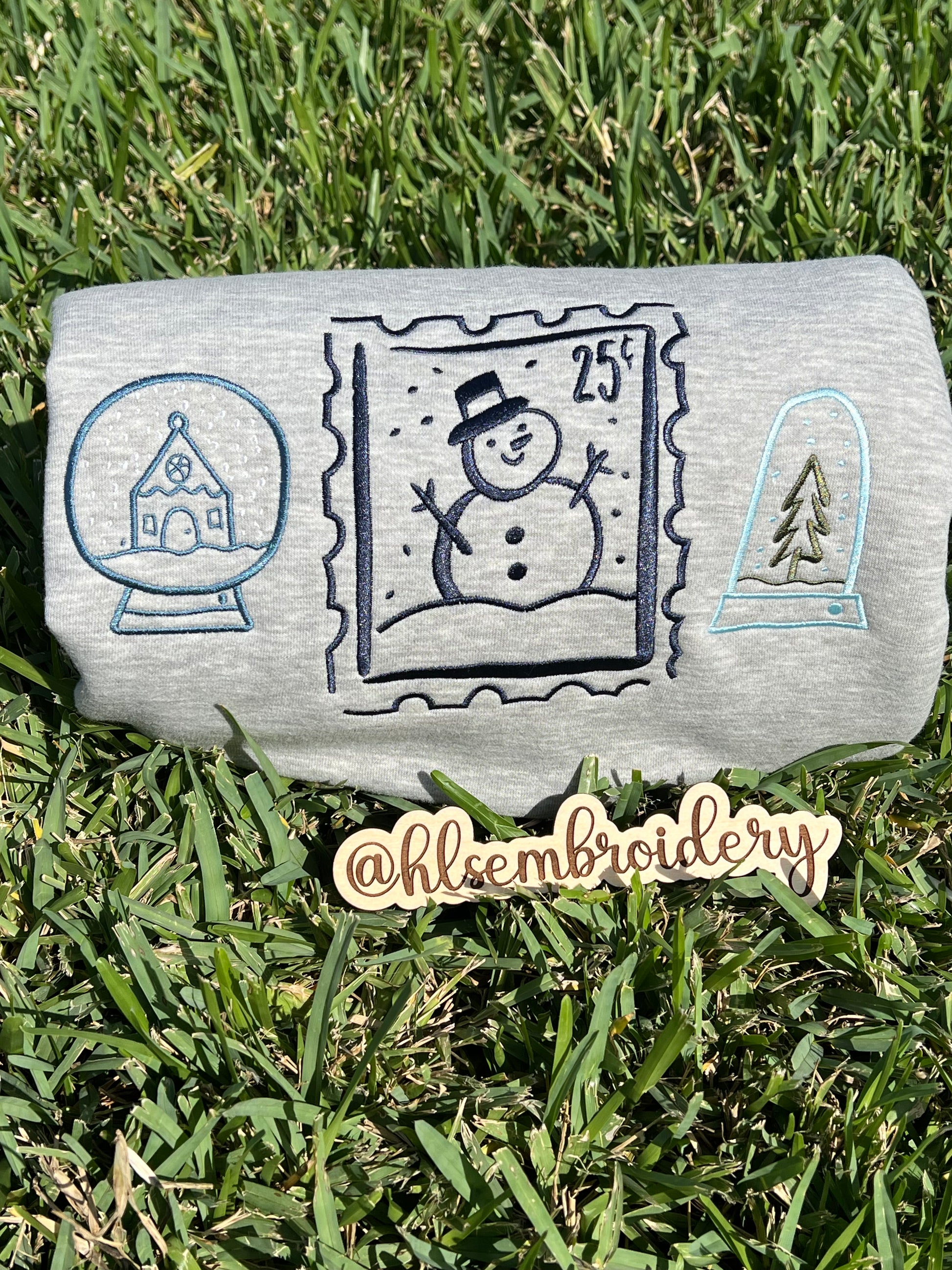 Embroidered designs of a snowman, snow globe, and pine tree on grass.