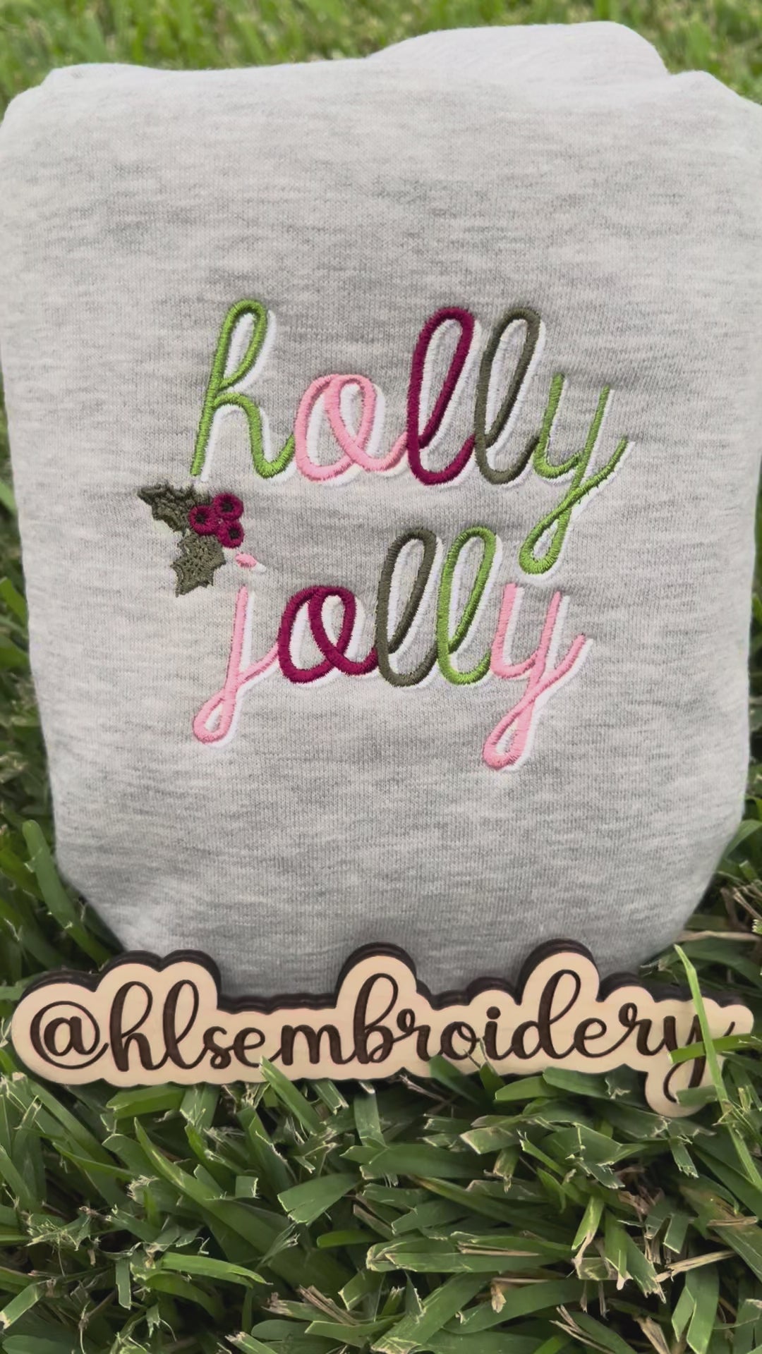 Folded gray color sweatshirt with 'holly jolly' embroidery on grass 